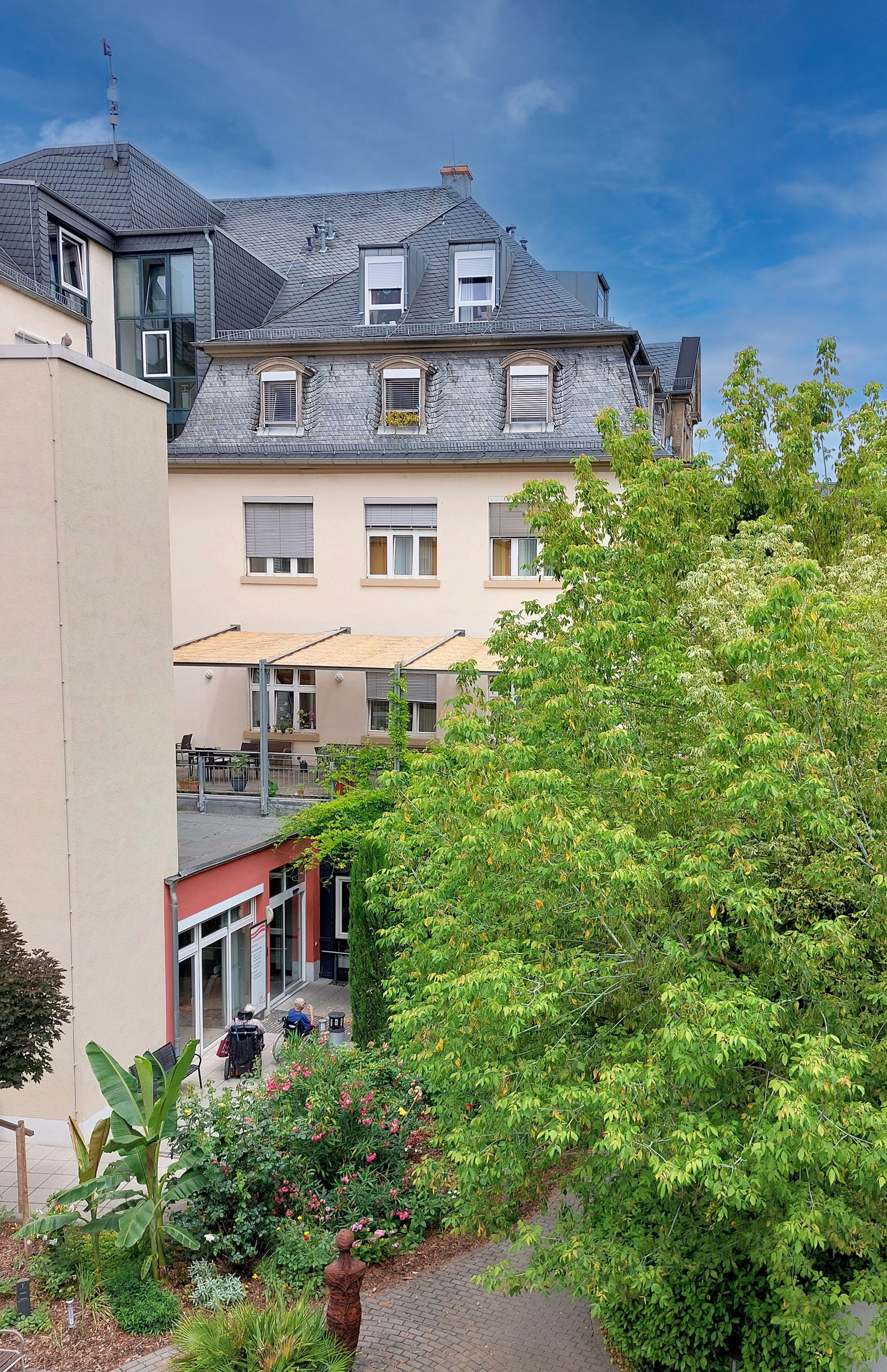 Blick auf das Gebäude des Burkhardhauses in Worms, das sich unter blauem Himmel befindet. Im Vordergrund ist ein grüner Baum zu sehen, der das Bild teilweise verdeckt. Auf der rechten Seite des Gebäudes ist eine Terrasse mit einem Sonnenschutzdach, Pflanzen und Tischen zu erkennen. Zwei Personen sitzen auf der Terrasse, während der Weg zum Eingang des Gebäudes von einem gepflasterten Weg gesäumt ist.