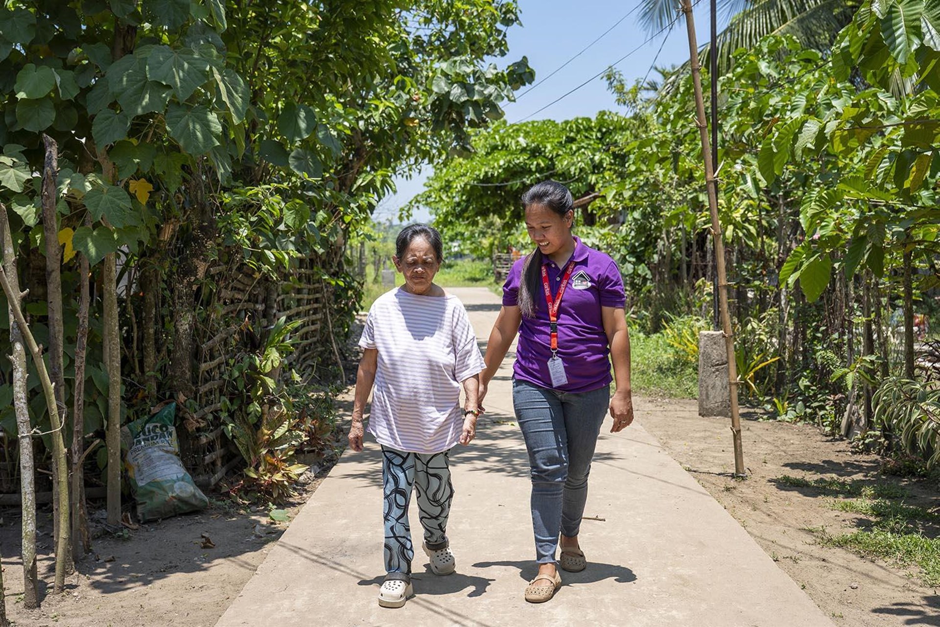 Das Bild zeigt eine kleinere ältere Frau und eine größere jüngere Frau, die auf einem Weg durch eine Plantage auf den Philippinen laufen.