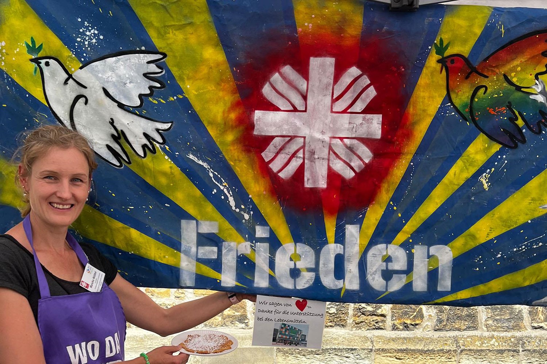 Eine Frau steht vor einer Fahne, auf der das Caritas-Logo, zwei Tauben und das Wort Frieden abgebildet sind.