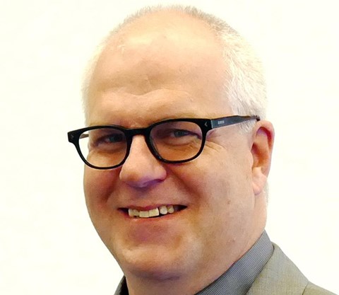 Lächelnder Mann mit weißen Haaren, Halbglatze und schwarzer Brille, der ein dunkelgraues Hemd und eine hellgraue Anzugjacke trägt