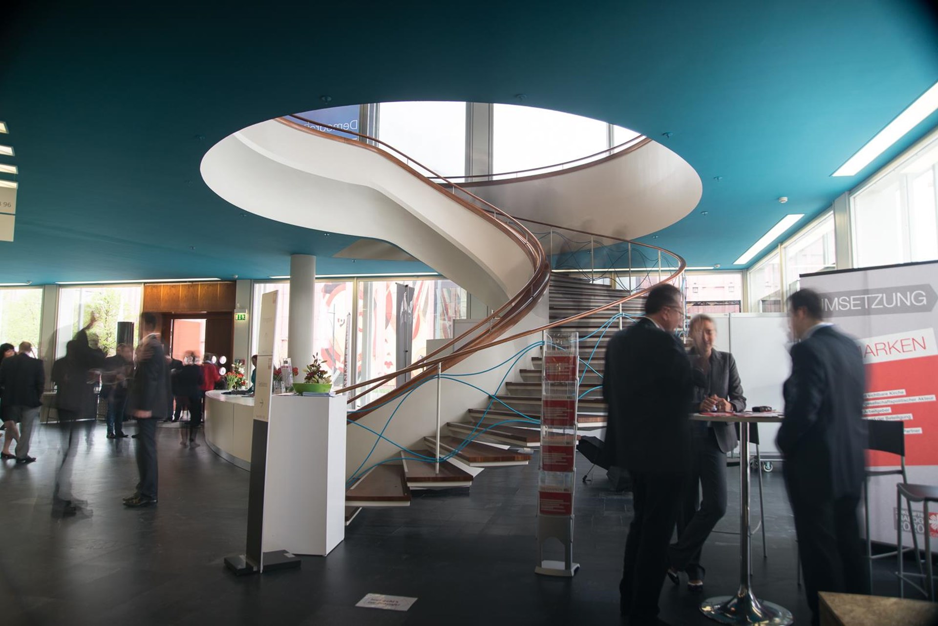 Moderne Treppe in einem hellen Foyer mit Geschäftsleuten im Gespräch. Der Raum hat große Fenster und eine auffällige Decke.