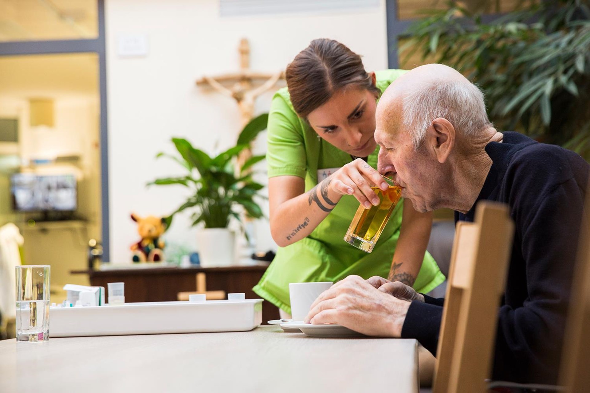 Pflegekraft hilft älterem Mann beim Trinken eines Glases Saft in einem hellen Raum mit Pflanzen.