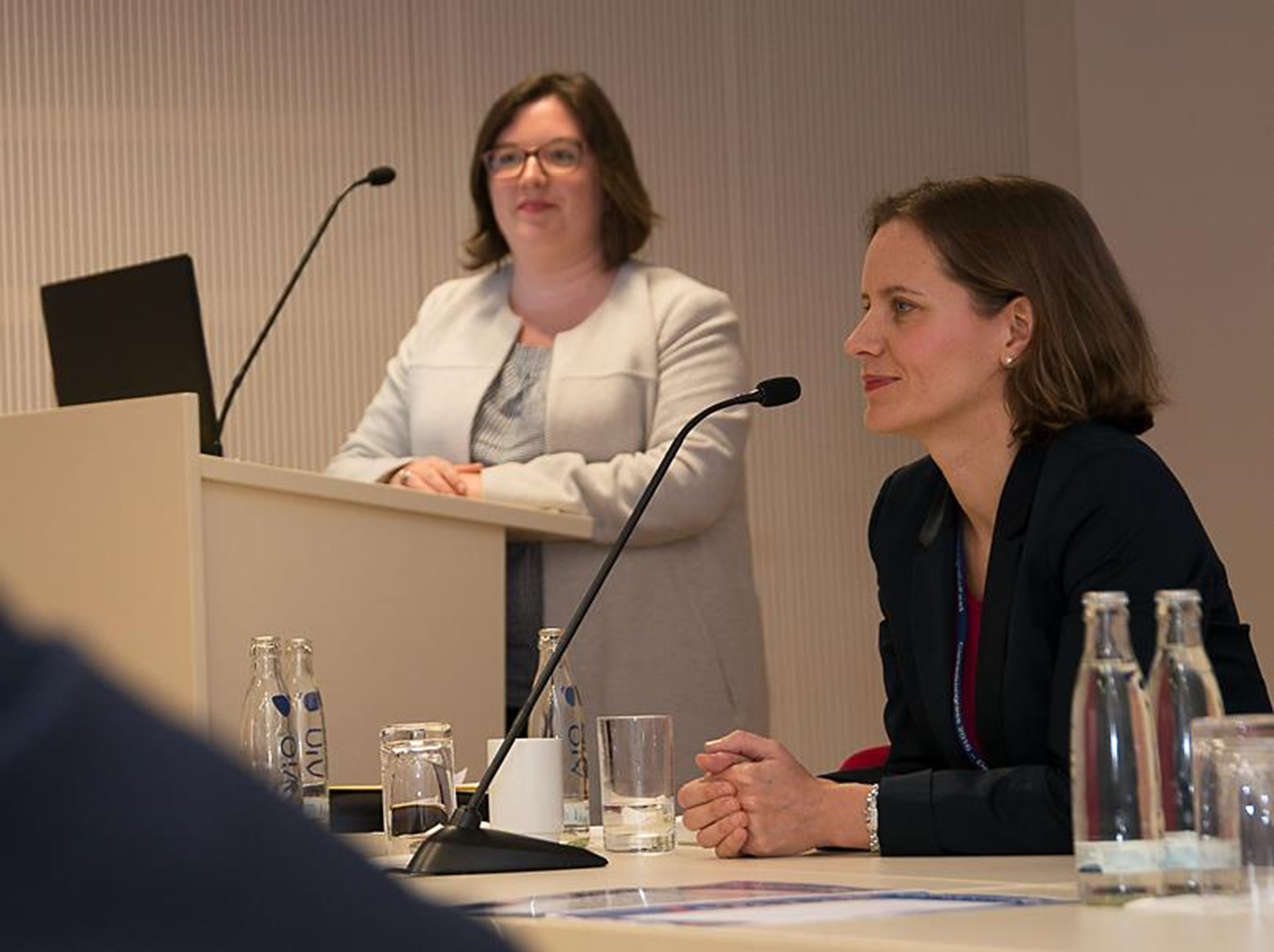 Zwei Frauen bei einer Konferenz. Eine Frau steht am Rednerpult, die andere sitzt und hört aufmerksam zu.