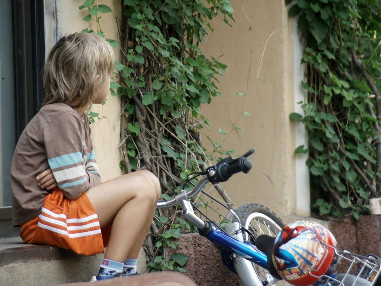 Ein Junge mit blonden, lockigen Haaren sitzt auf einer Treppe, gekleidet in ein braunes Shirt und orangefarbene Shorts, während er nachdenklich in die Ferne schaut. 