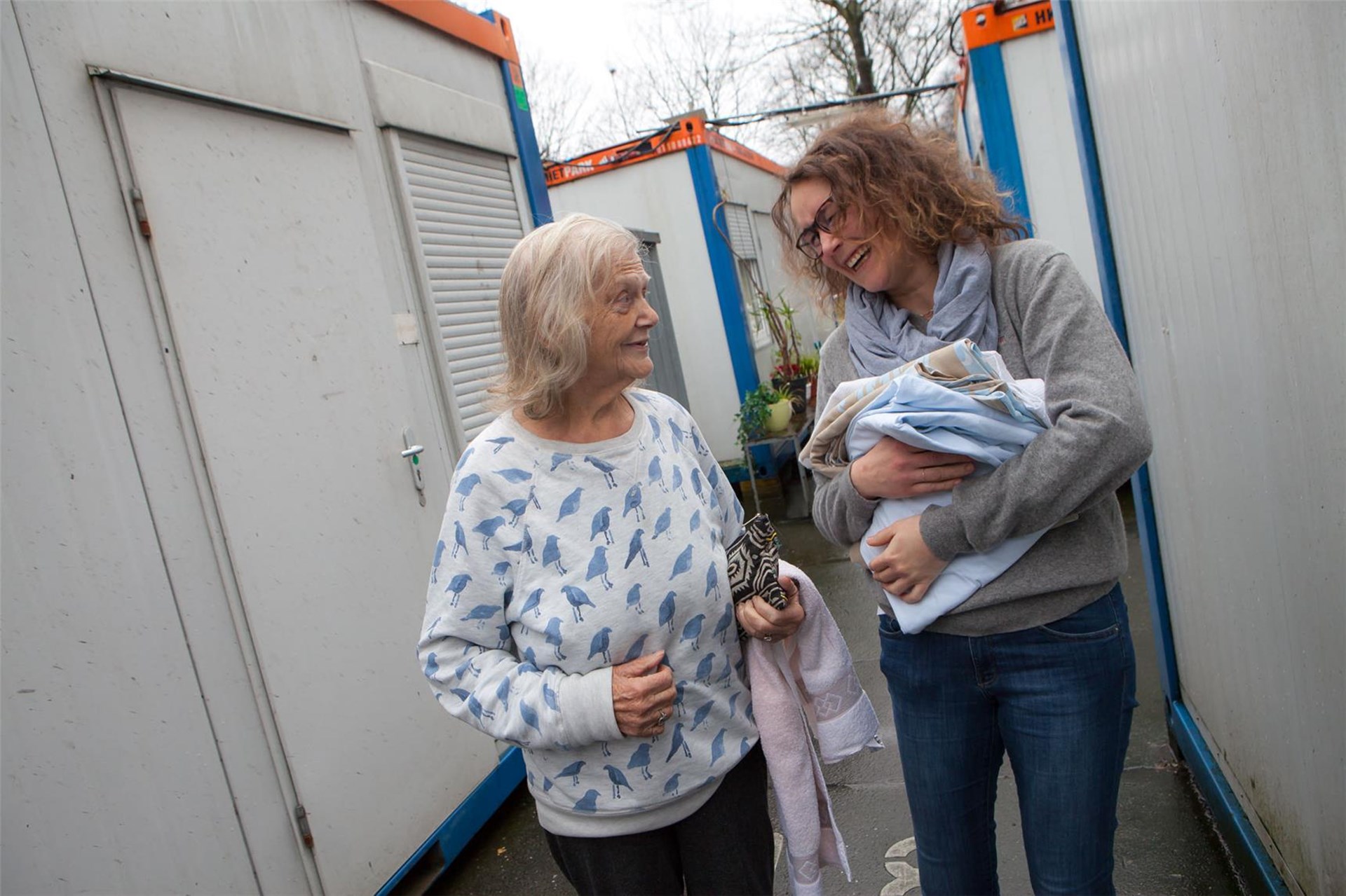 Zwei Frauen unterschiedlichen Alters unterhalten sich draußen zwischen Containern, eine hält ein Baby und die andere trägt eine Jacke.