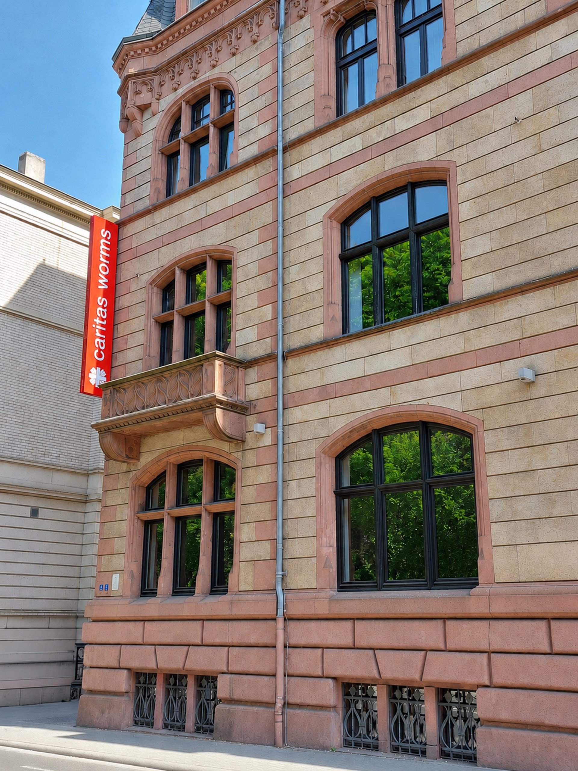 Straßenansicht eines mehrstöckigen historischen Gebäudes mit hell beiger und rosafarbener Sandsteinfassade und Rundbogenfenstern. An der linken Seite hängt ein vertikales rotes Schild mit der Aufschrift „caritas worms“. In den Fenstern spiegeln sich grüne Bäume; das Gebäude steht in einer städtischen Umgebung bei Sonnenschein.