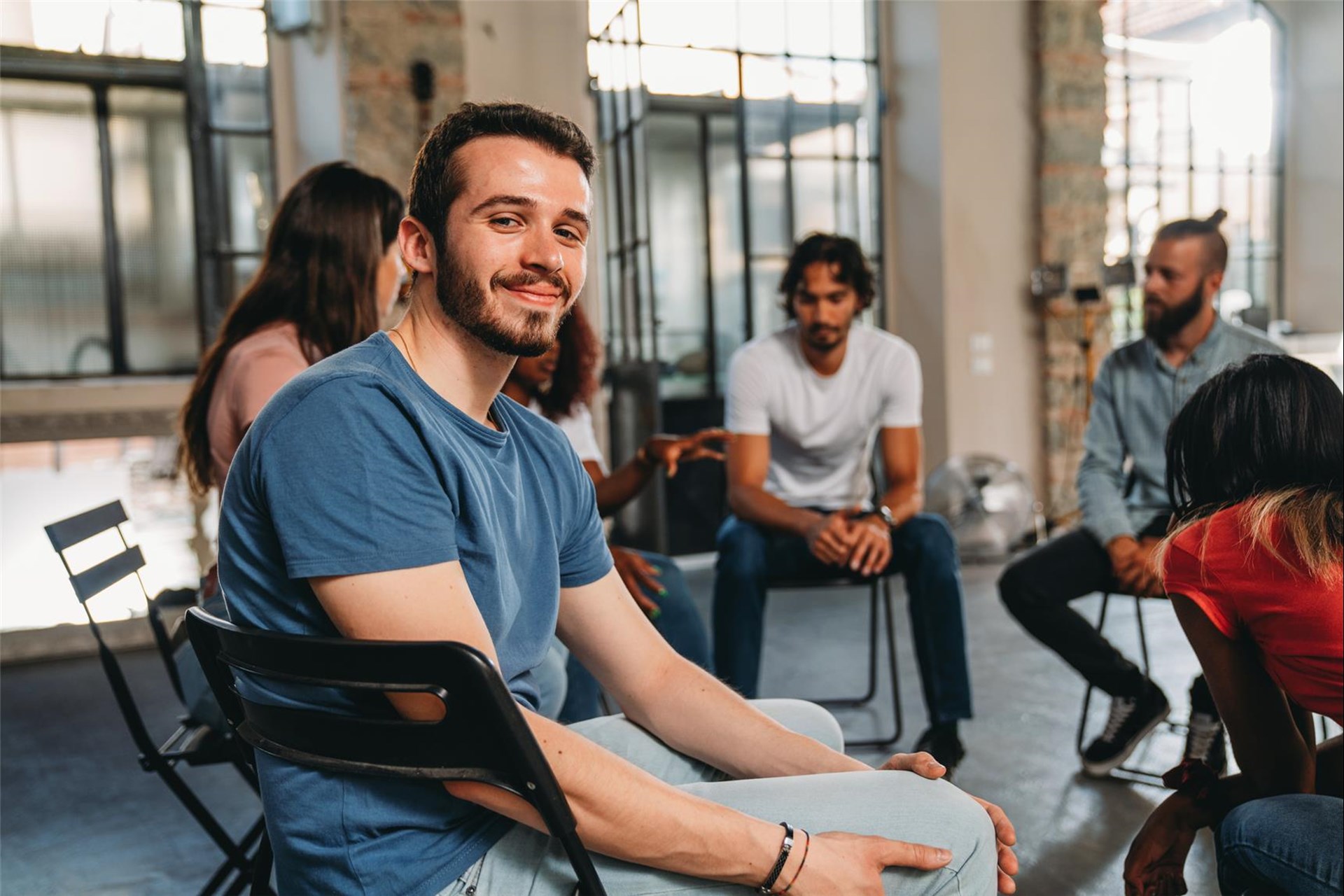 Junger Mann mit Bart sitzt auf einem Stuhl und lächelt, während er Teil einer Gruppenrunde in einem modernen Raum ist.