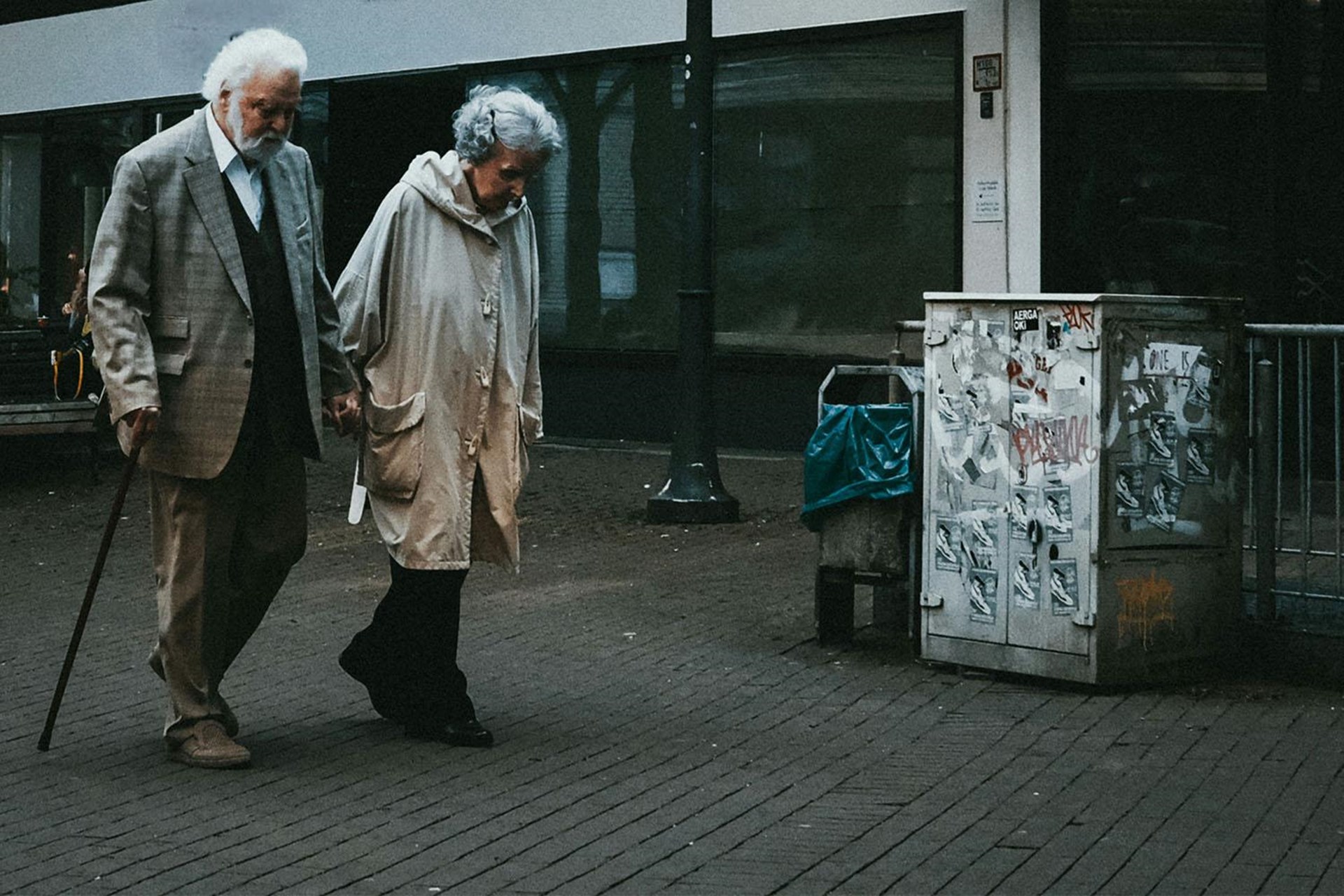 Ein alten Paar geht gebeugt und Hand (er mit Stock) in Hand einen tristen Platz entlang.