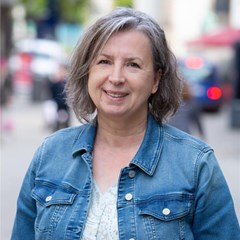 Lächelnde Frau mit grauen Haaren, die ein helles T-Shirt mit einer blauen Jeansjacke trägt und sich in der Fußgängerzone der Essener Innenstadt befindet