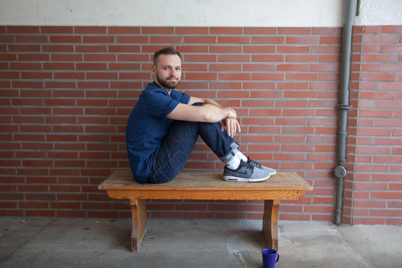 Mann sitzt auf einer Holzbank, umarmt seine Knie, trägt ein blaues Shirt und Jeans, mit Sneakers. Hintergrund aus roten Ziegelsteinen.