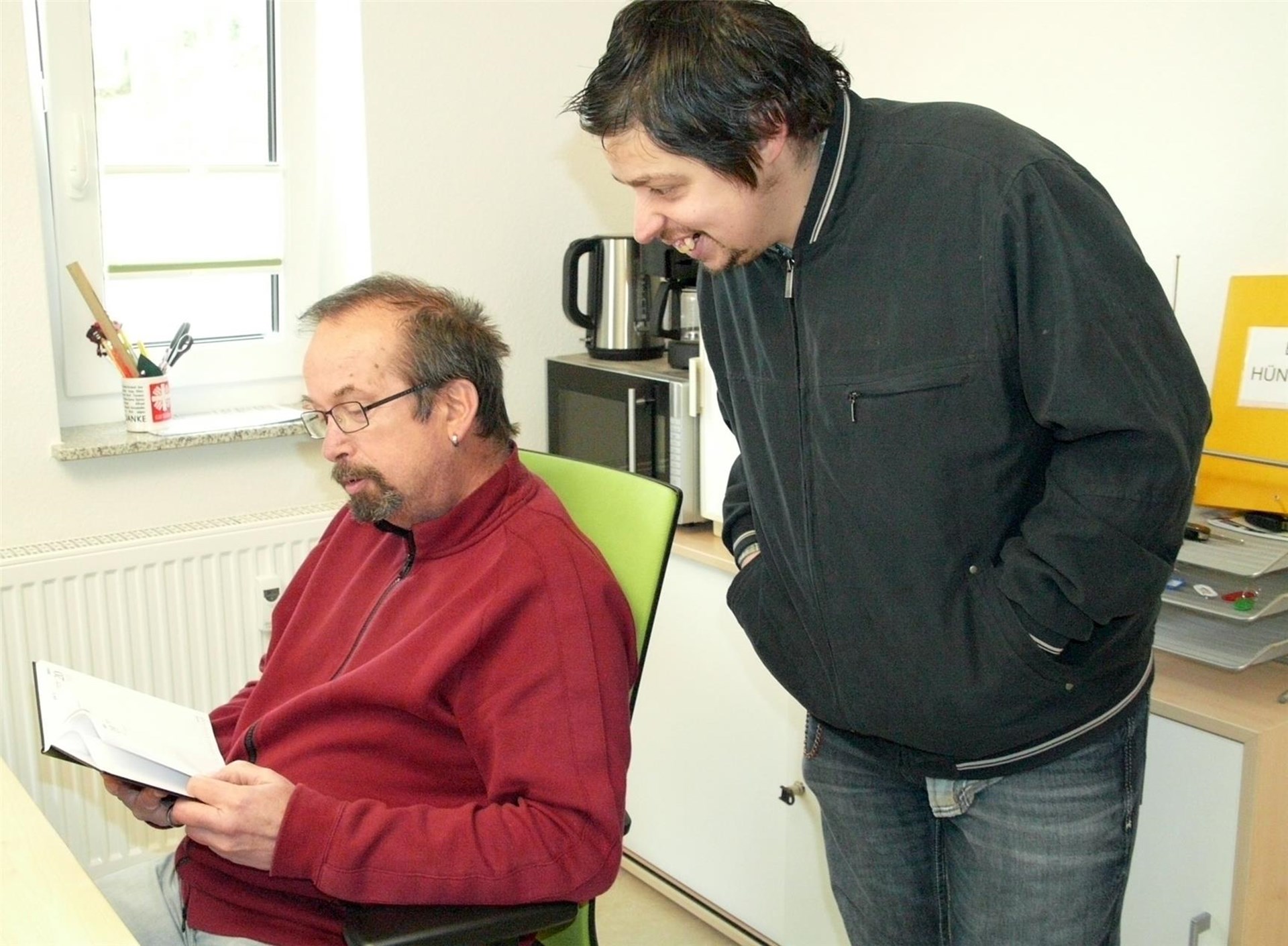 Zwei Männer in einem Büro, einer sitzt und liest ein Dokument, der andere steht und schaut interessiert darauf.