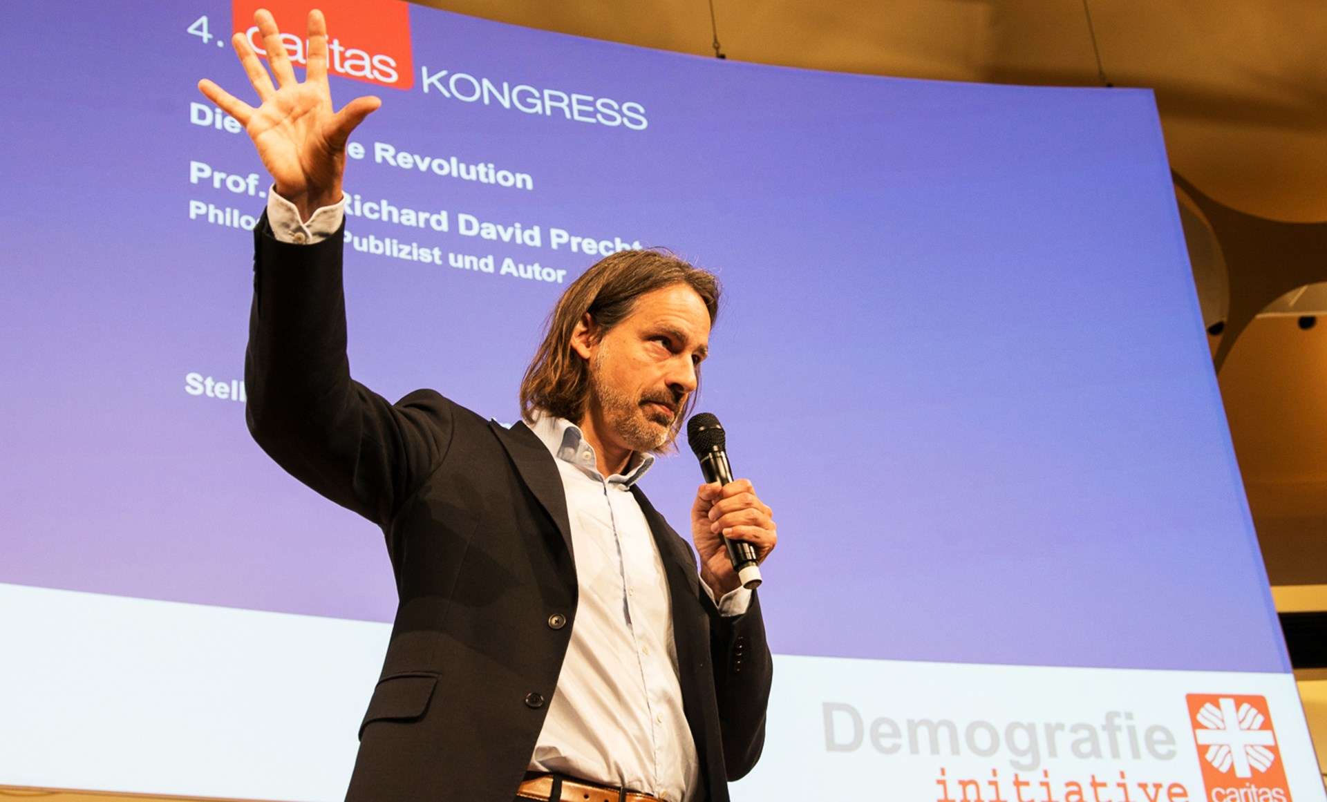 Richard David Precht spricht auf dem 4. Congress der Initiative Demografie, hält ein Mikrofon und gestikuliert.