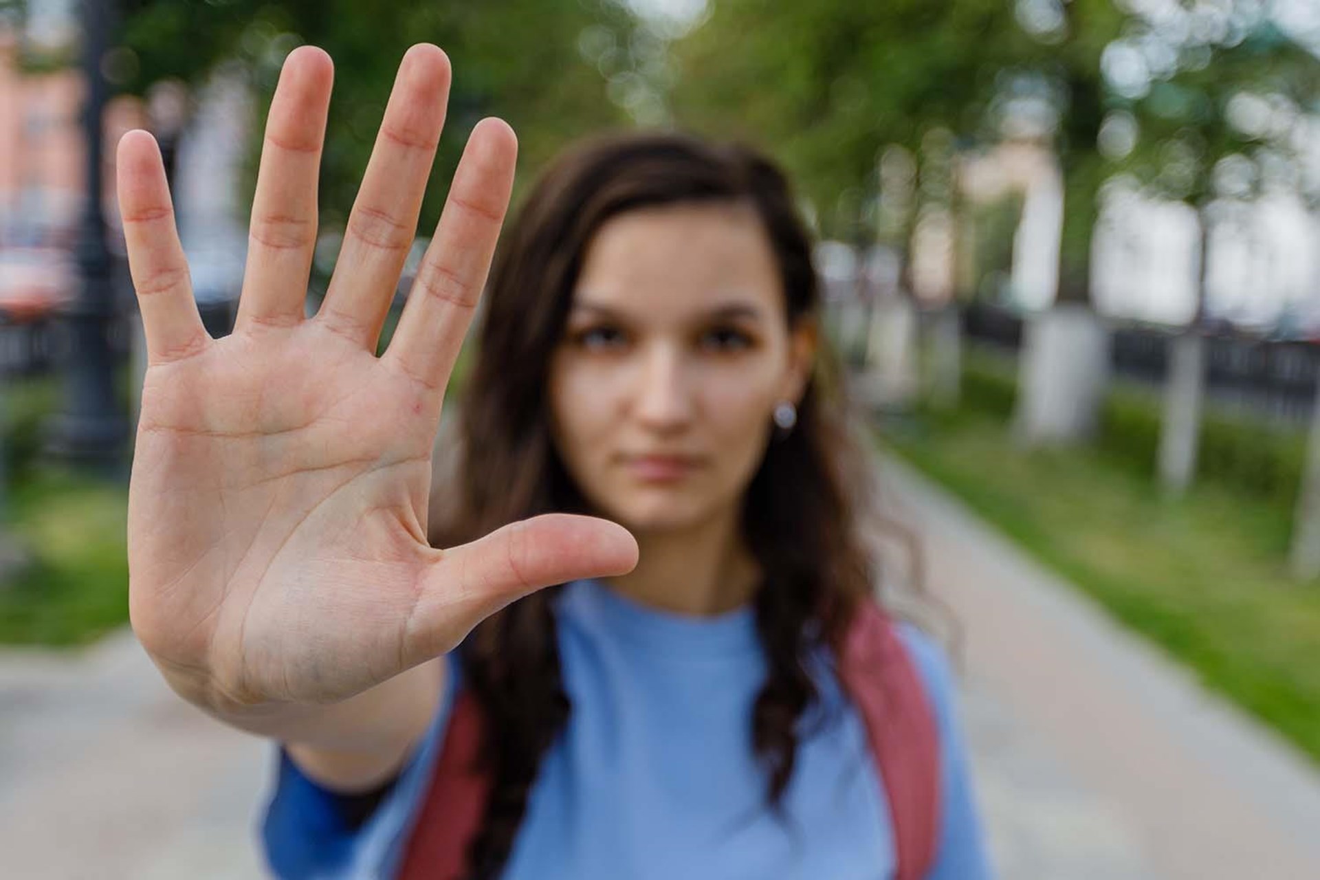 Junge Frau mit langen, lockigen Haaren, die die Hand in einer gestoppten Pose nach vorne zeigt. Sie trägt ein blaues Oberteil und hat einen Rucksack auf.