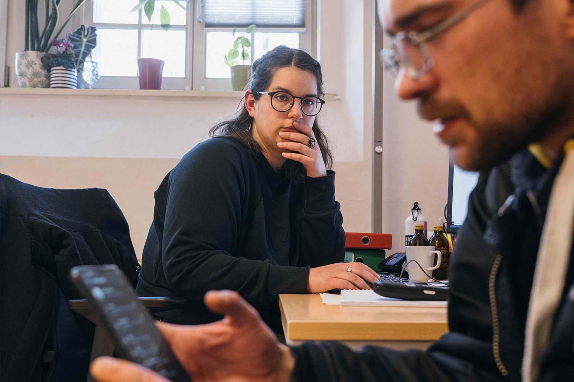 Eine junge Frau, im schwarz bekleidet sitzt denkend am Schreibtisch. 