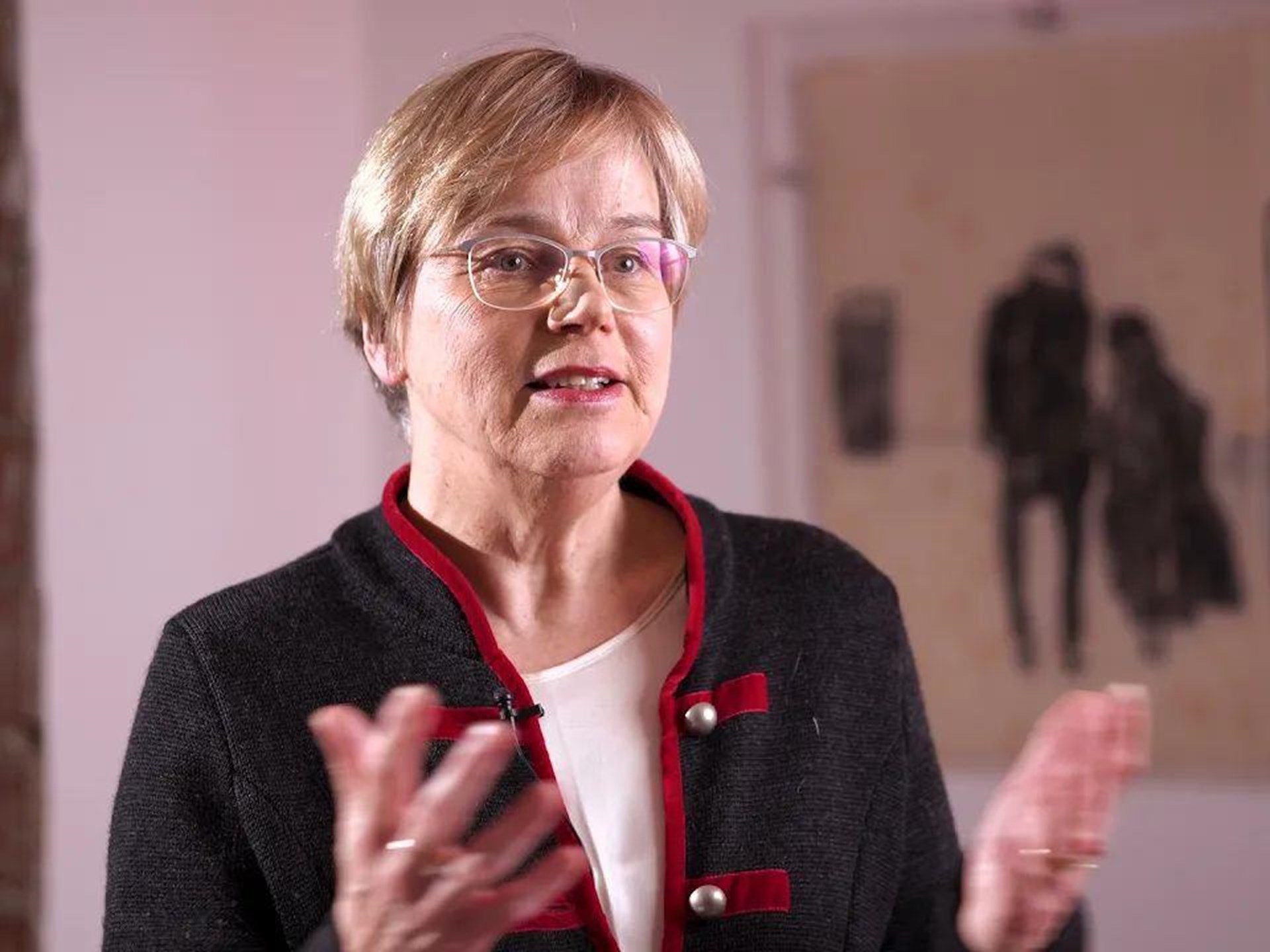 Frau mit blonden, kurzen Haaren und Brille, trägt einen schwarzen Blazer mit roten Akzenten. Sie spricht und gestikuliert, im Hintergrund sind verschwommene Kunstwerke zu sehen.