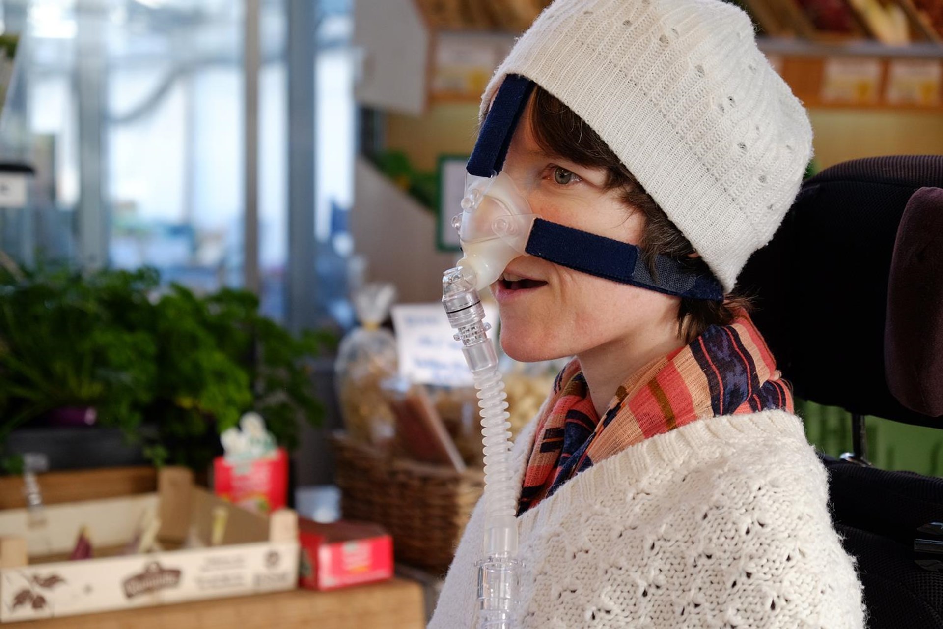 Frau mit Atemmaske sitzt in einem Geschäft, trägt eine gestrickte Mütze und ein weißes Oberteil. Im Hintergrund sind frische Lebensmittel zu sehen.