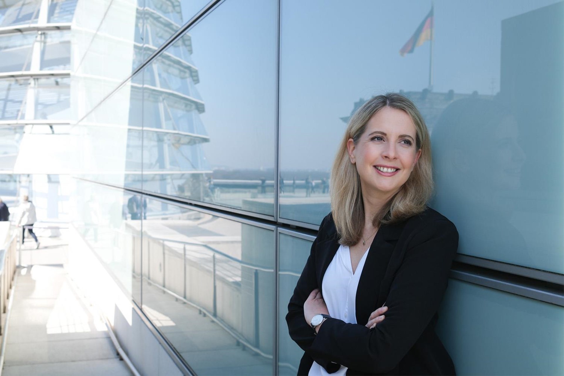 Bundesbauministerin Verena Hubertz (SPD), eine Frau mit langen blonden Haaren, lehnt an einer Glasfassade, in der sie sich spiegelt. Im Hintergrund sieht man die Spiegelung eines Teils des Bundestags mit der gehissten Deutschlandflagge. 