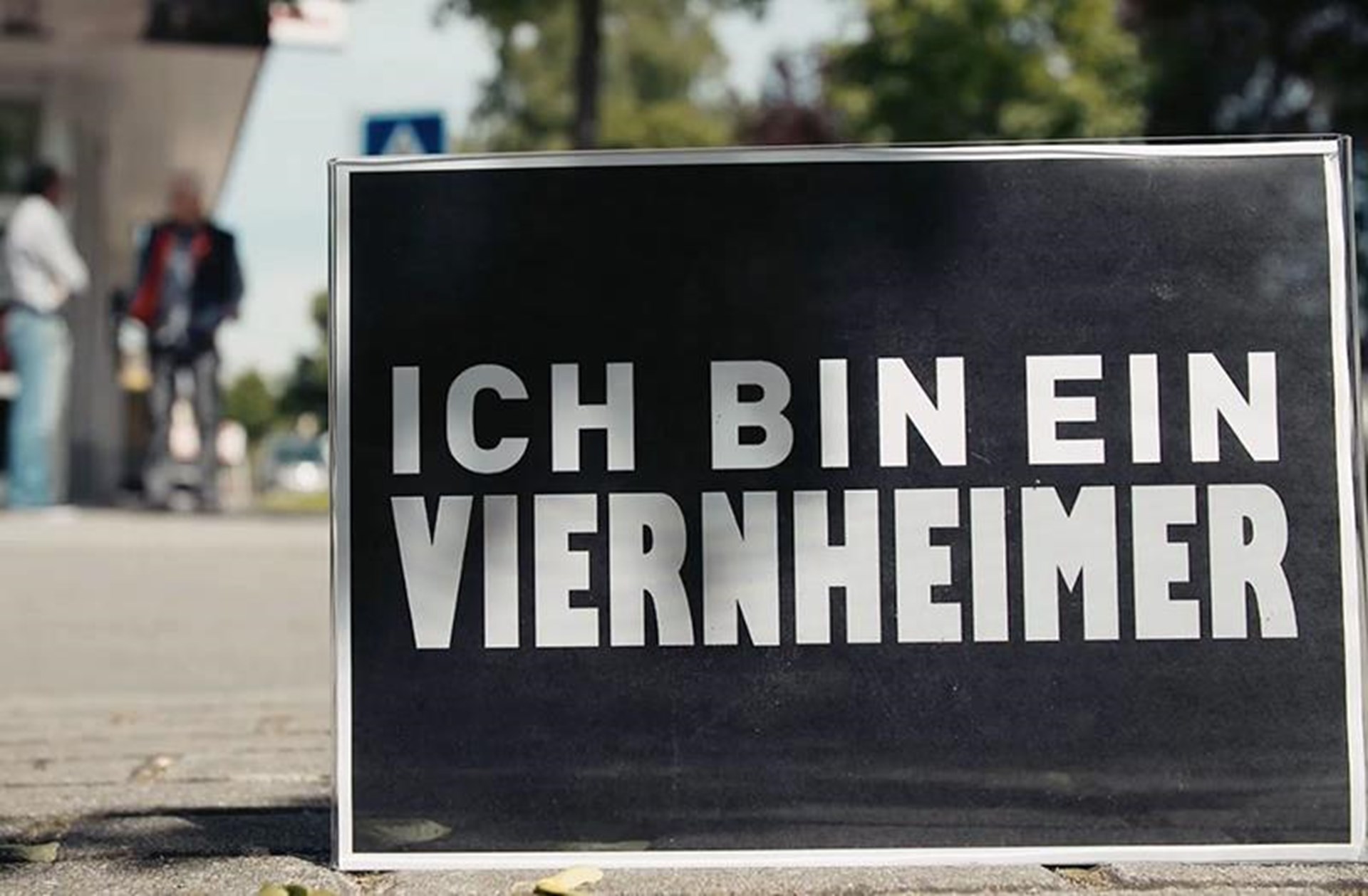 Schwarzes Schild mit weißem Text "ICH BIN EIN VIERNHEIMER" auf einem Gehweg, im Hintergrund unscharfe Personen.