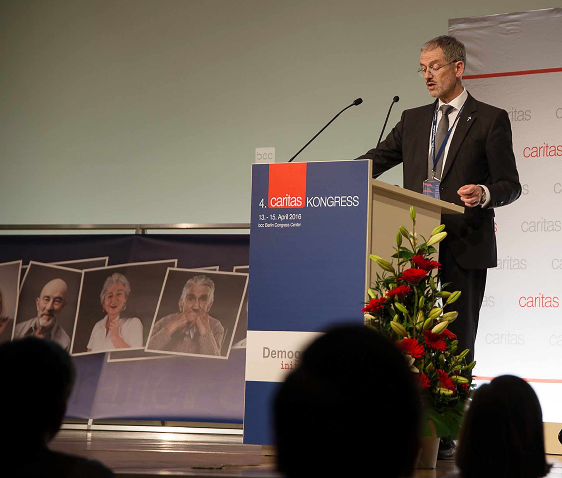 Mann am Rednerpult beim Caritas-Kongress 2016, im Hintergrund Porträts von Teilnehmern.