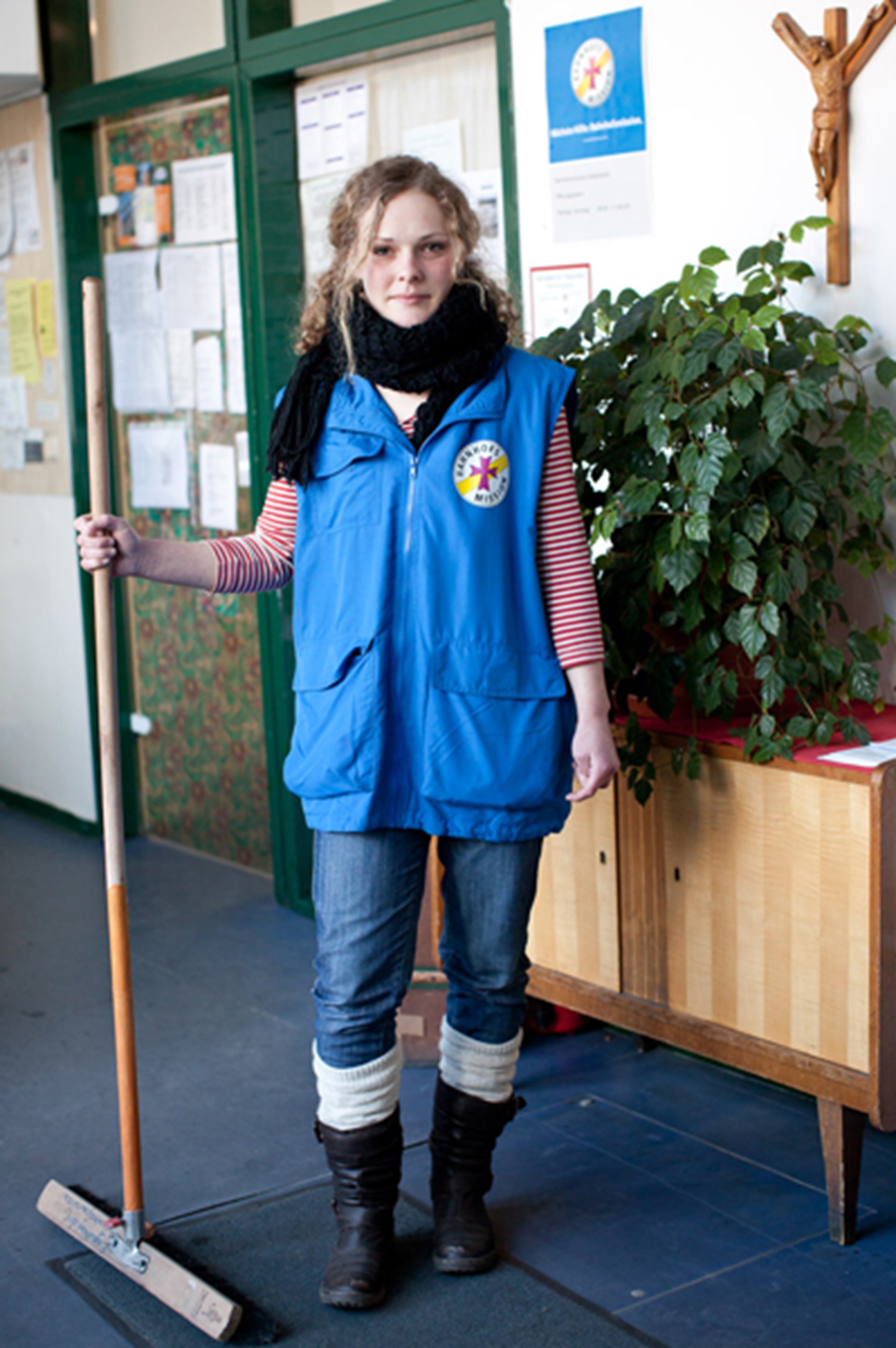 Junge Frau in blauer Weste mit Logo, gestreiftem Shirt und Jeans, hält einen Besen in der Hand. Hintergrund zeigt eine Pflanze und eine Wand mit Dokumenten.