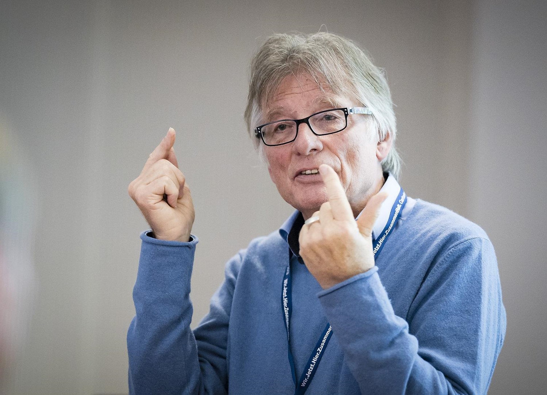 Mann mit grauen Haaren und Brille, trägt einen blauen Pullover, spricht mit erhobenen Händen und gestikuliert.