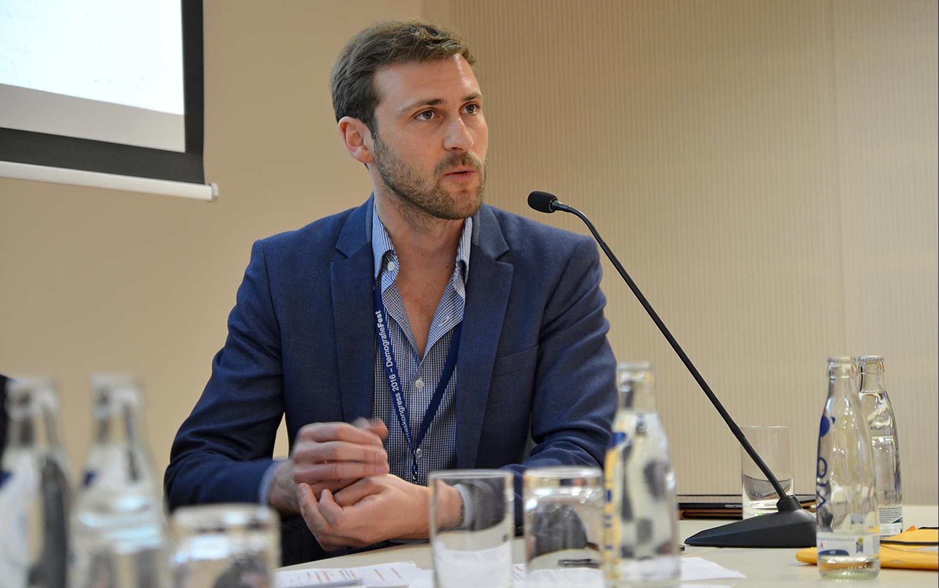 Mann in blauer Jacke spricht an einem Tisch mit Wasserflaschen und Mikrofon während einer Konferenz.