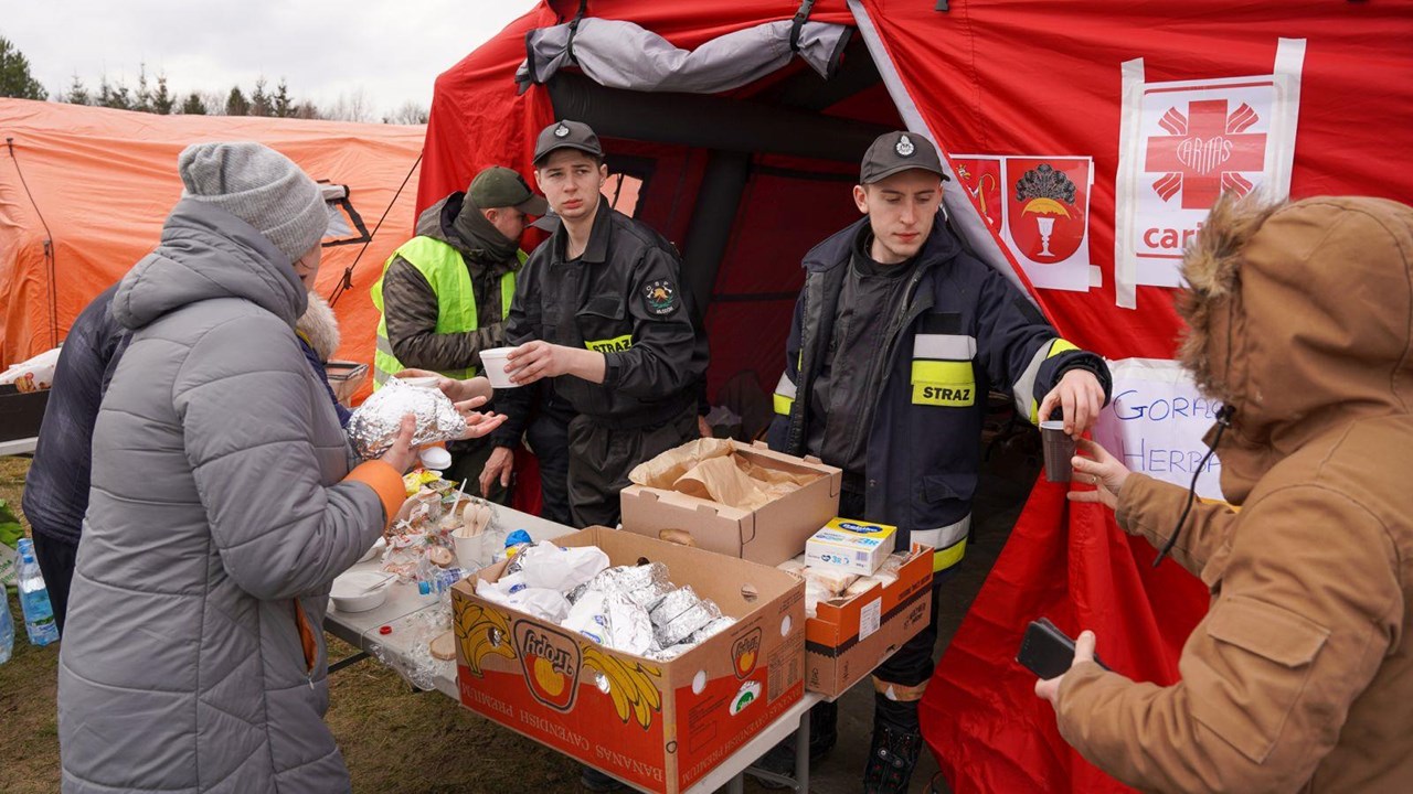 Zwei Helfer in Uniform verteilen Essen an Bedürftige vor einem roten Zelt, das mit einem Hilfslogo versehen ist.
