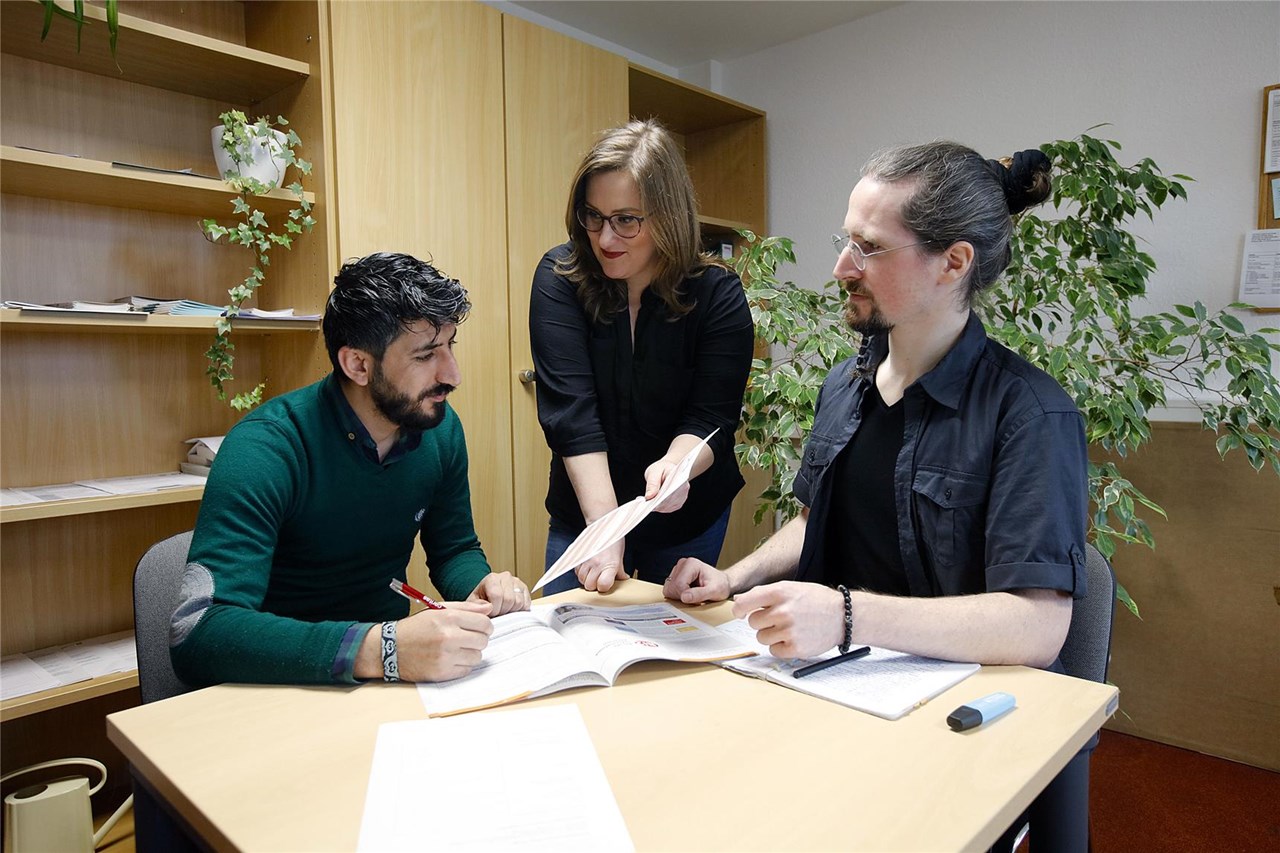 Drei Personen sitzen an einem Tisch und arbeiten zusammen. Ein Mann mit dunklem Bart und grünem Pullover schreibt, während eine Frau mit Brille und blonden Haaren ihm ein Dokument zeigt.