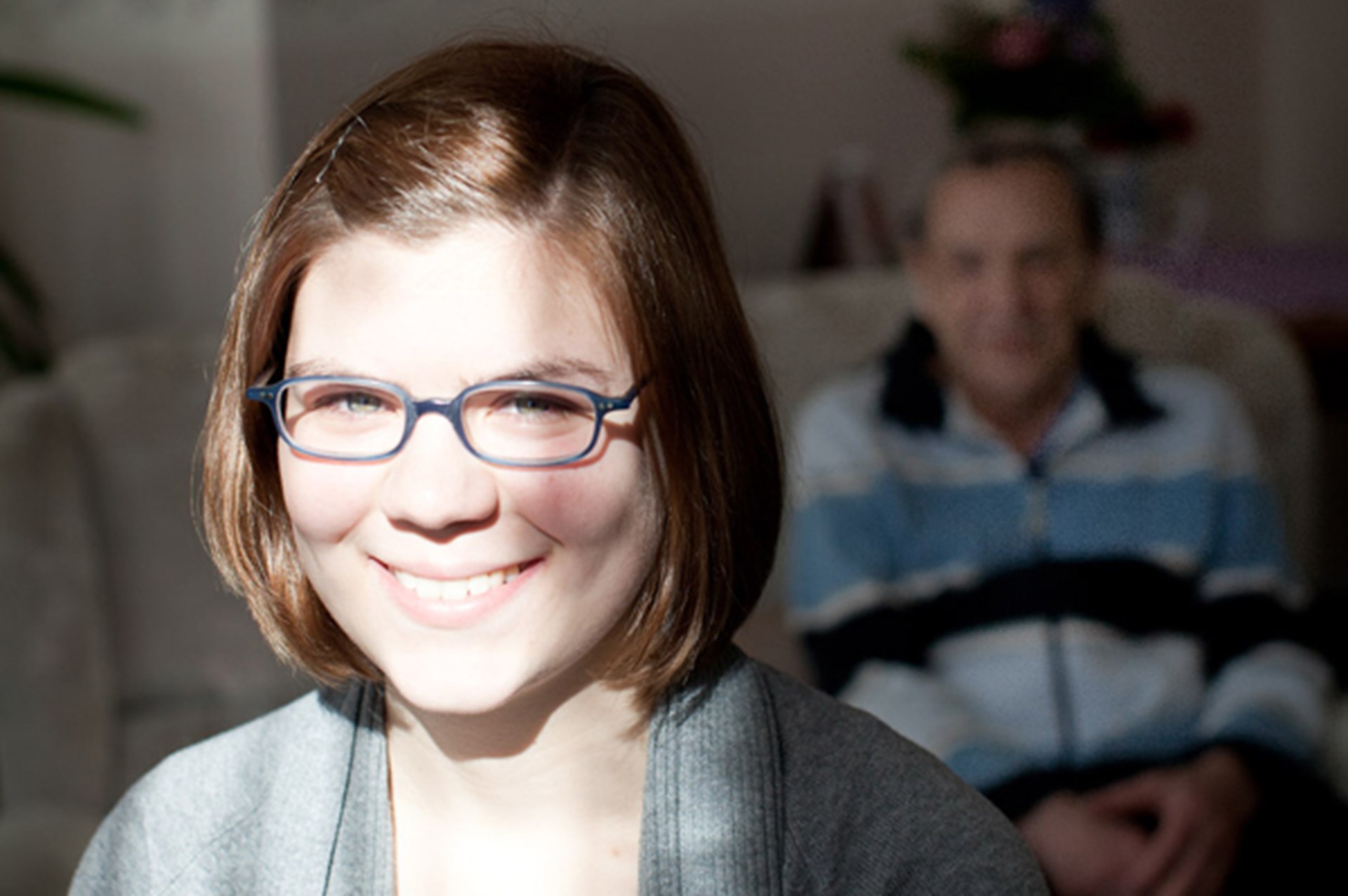Lächelnde junge Frau mit Brille und kurzem Haar, im Hintergrund sitzt ein älterer Mann in einem blauen gestreiften Pullover.