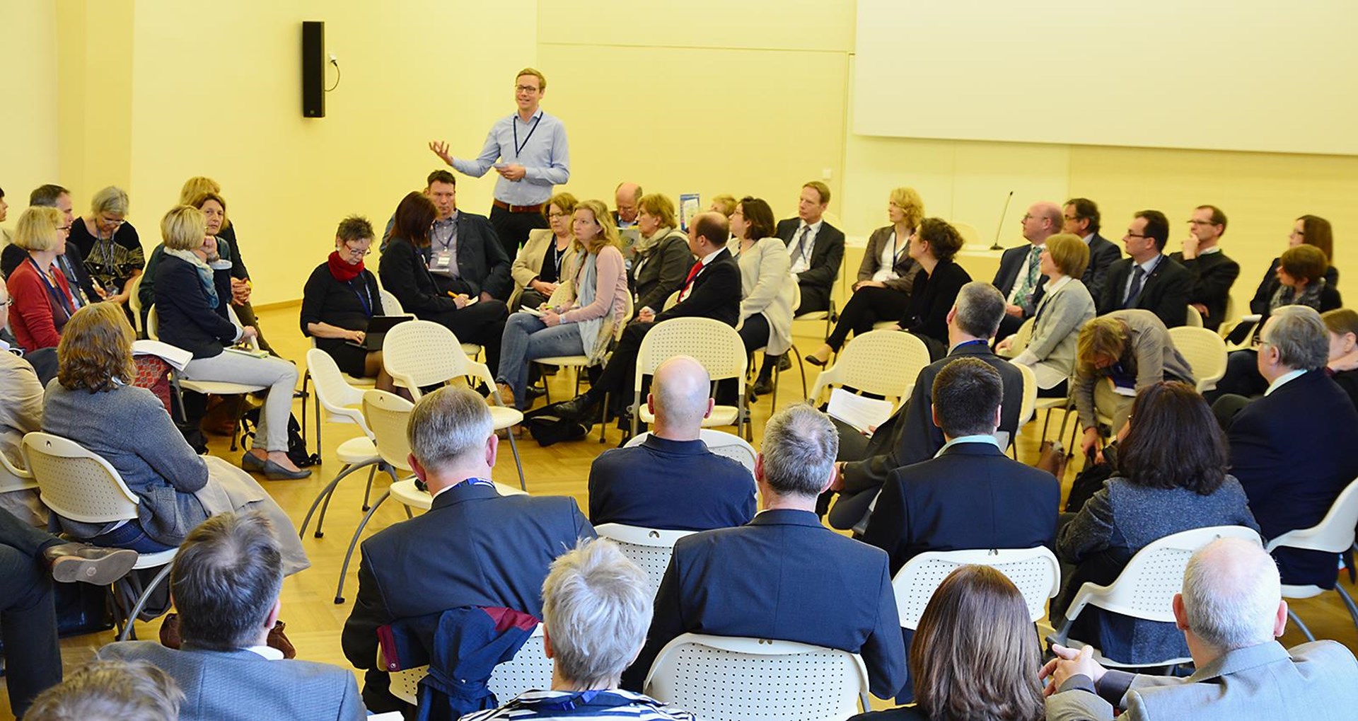 Eine Gruppe von Menschen in einem großen Raum, die in einem Kreis sitzen. Ein Mann steht in der Mitte und spricht zu den Anwesenden.