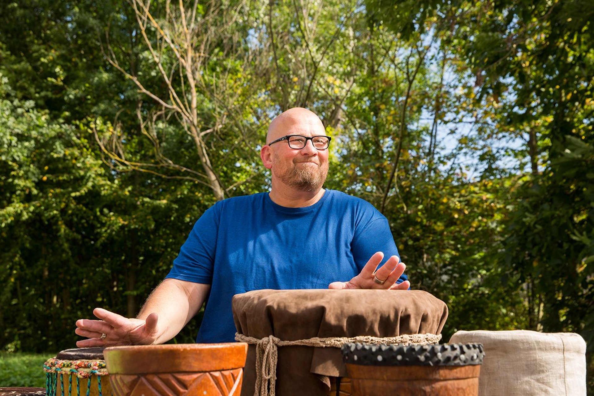 Mann mit Brille und blauem T-Shirt spielt draußen auf verschiedenen Trommeln, umgeben von Bäumen.