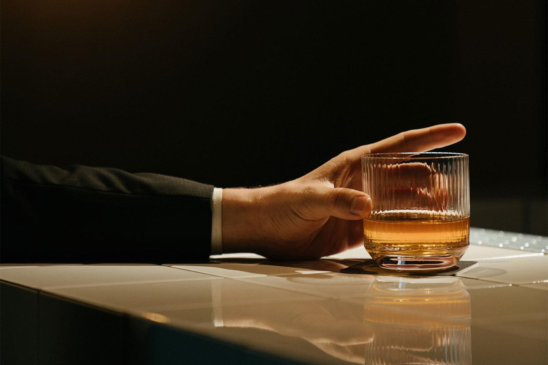 Eine Hand in einem Anzug hält ein Glas mit goldenem Getränk an einer Bar. Der Hintergrund ist dunkel, die Oberfläche glänzt.