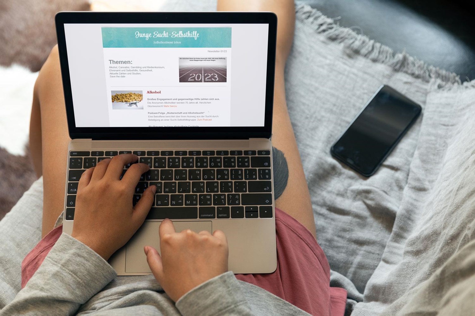 Person mit grauem Langarmshirt und rosa Shorts tippt auf einem Laptop, umgeben von einem grauen Sofa und einem Smartphone.