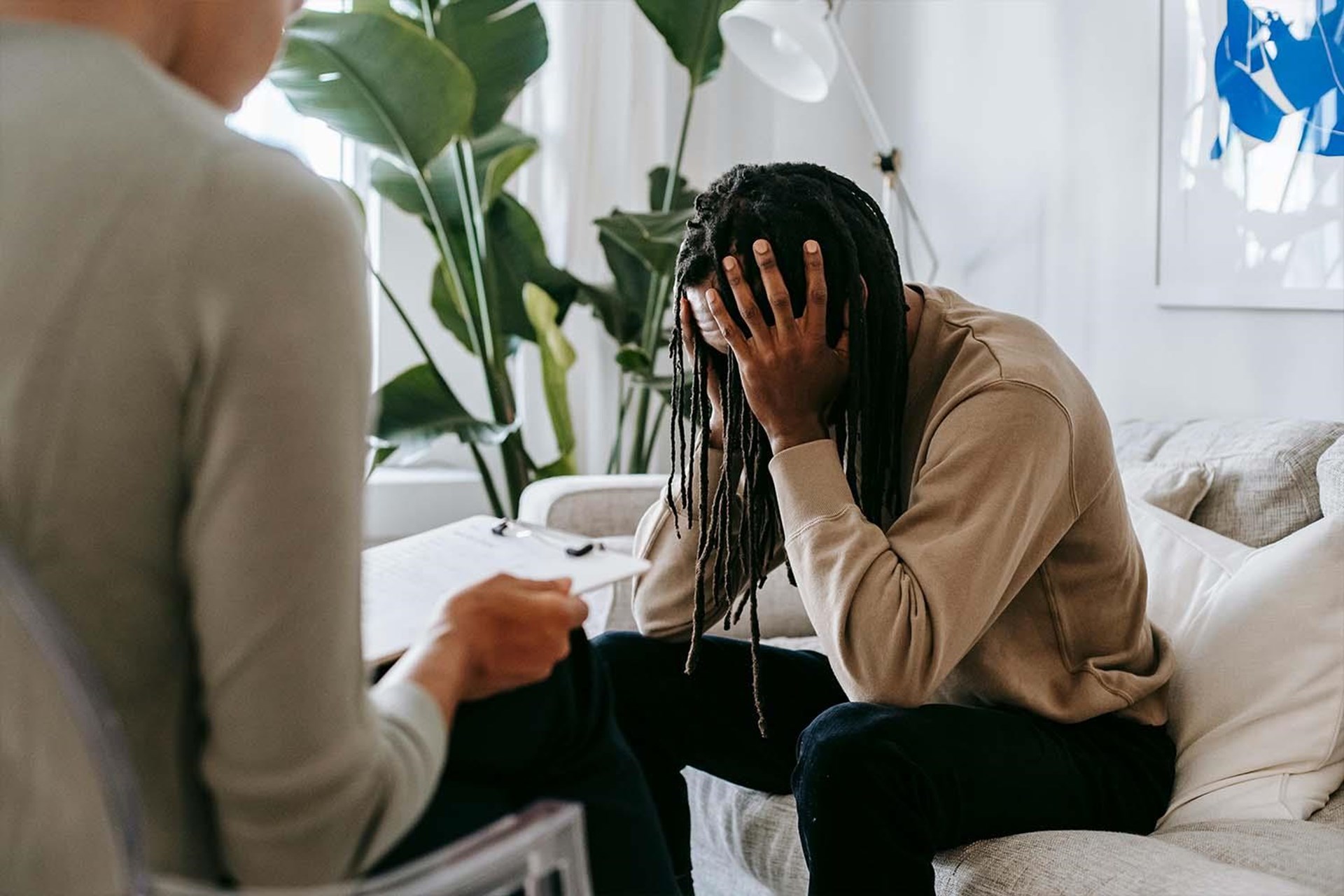 Ein Mann mit langen Haaren, der frustriert auf einem Sofa sitzt, hält den Kopf in den Händen. Eine Person sitzt ihm gegenüber, nicht sichtbar, in einem hellen Raum mit Pflanzen im Hintergrund.