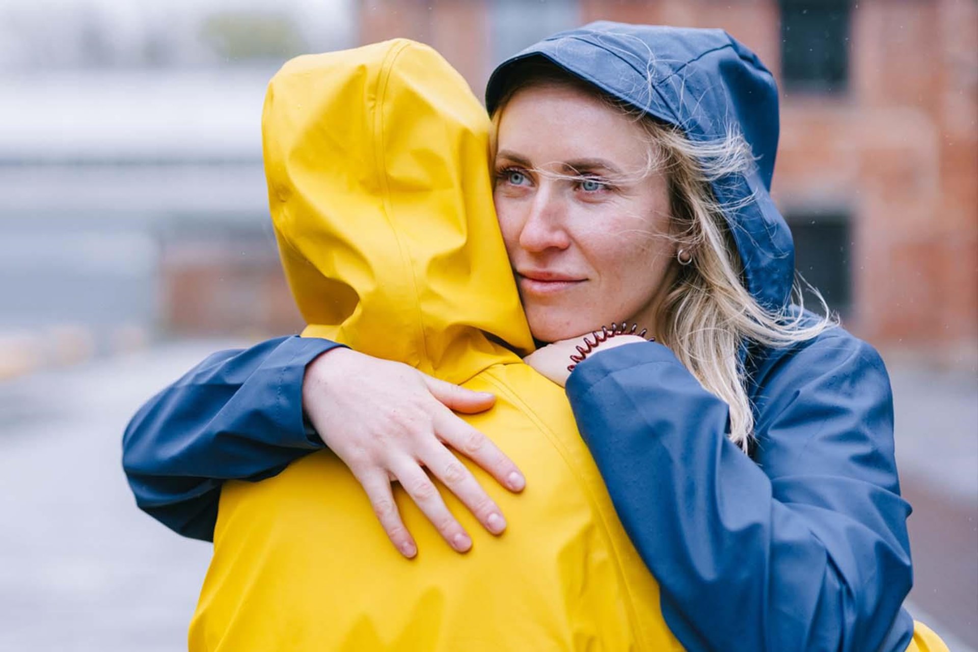 Frau mit blonden Haaren in blauer Regenjacke umarmt jemanden in gelber Regenjacke, zeigt emotionale Verbindung an einem regnerischen Tag.