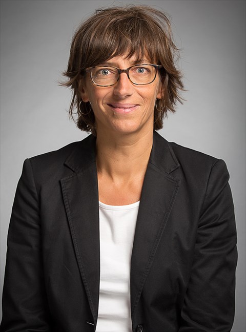 Lächelnde Frau mit braunen Haaren und Brille, die ein weißes T-Shirt und eine schwarze Anzugjacke trägt
