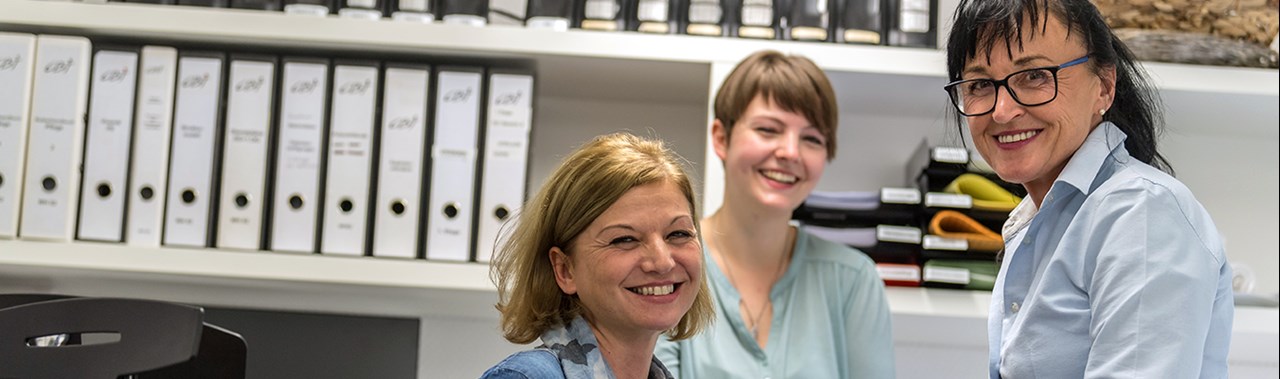 Drei Frauen in einem Büro lächeln in die Kamera. Im Hintergrund sind Aktenordner sichtbar.