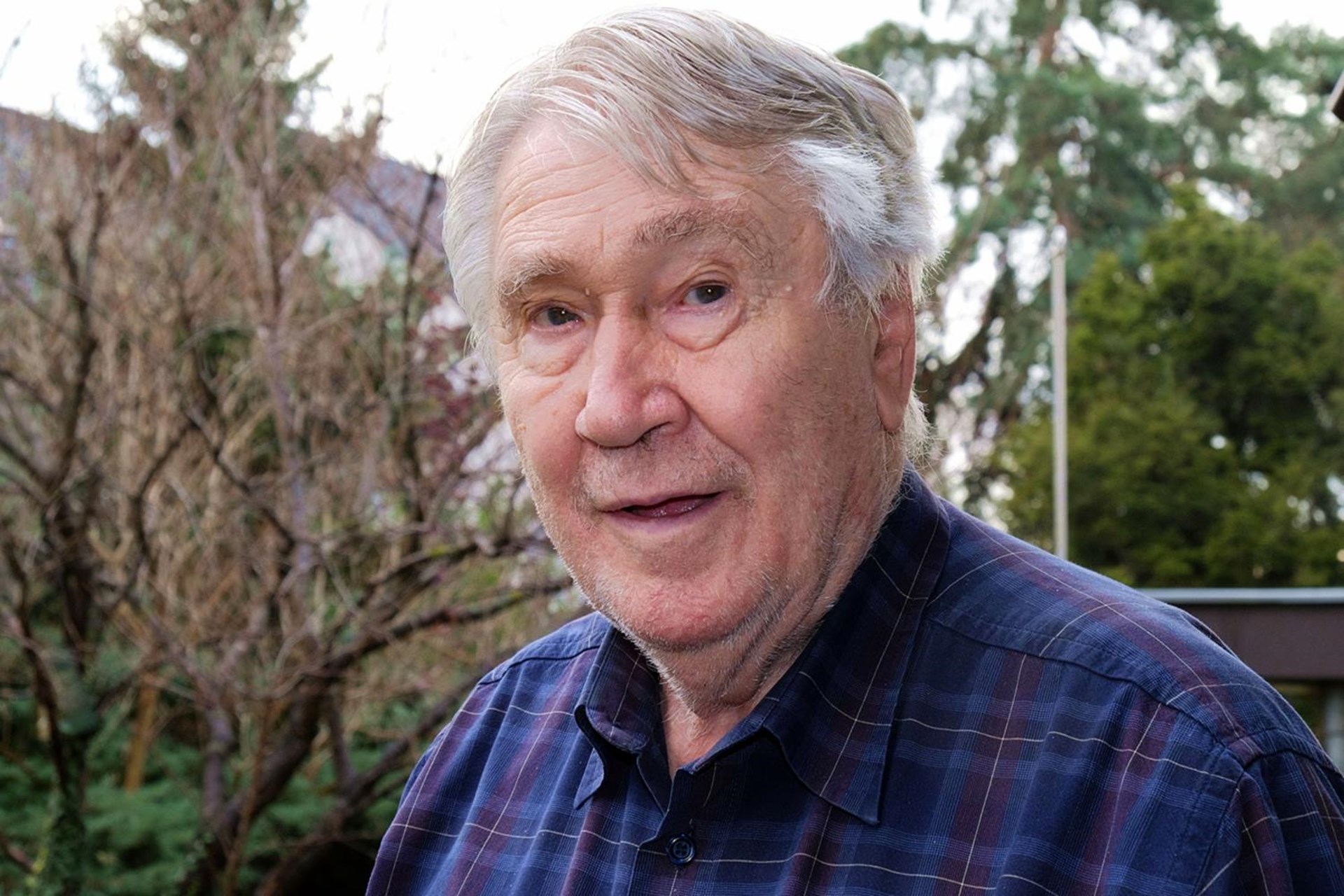 Älterer Mann mit grauen Haaren und freundlichem Gesichtsausdruck, trägt ein kariertes Hemd. Hintergrund mit Bäumen und Gebäuden.