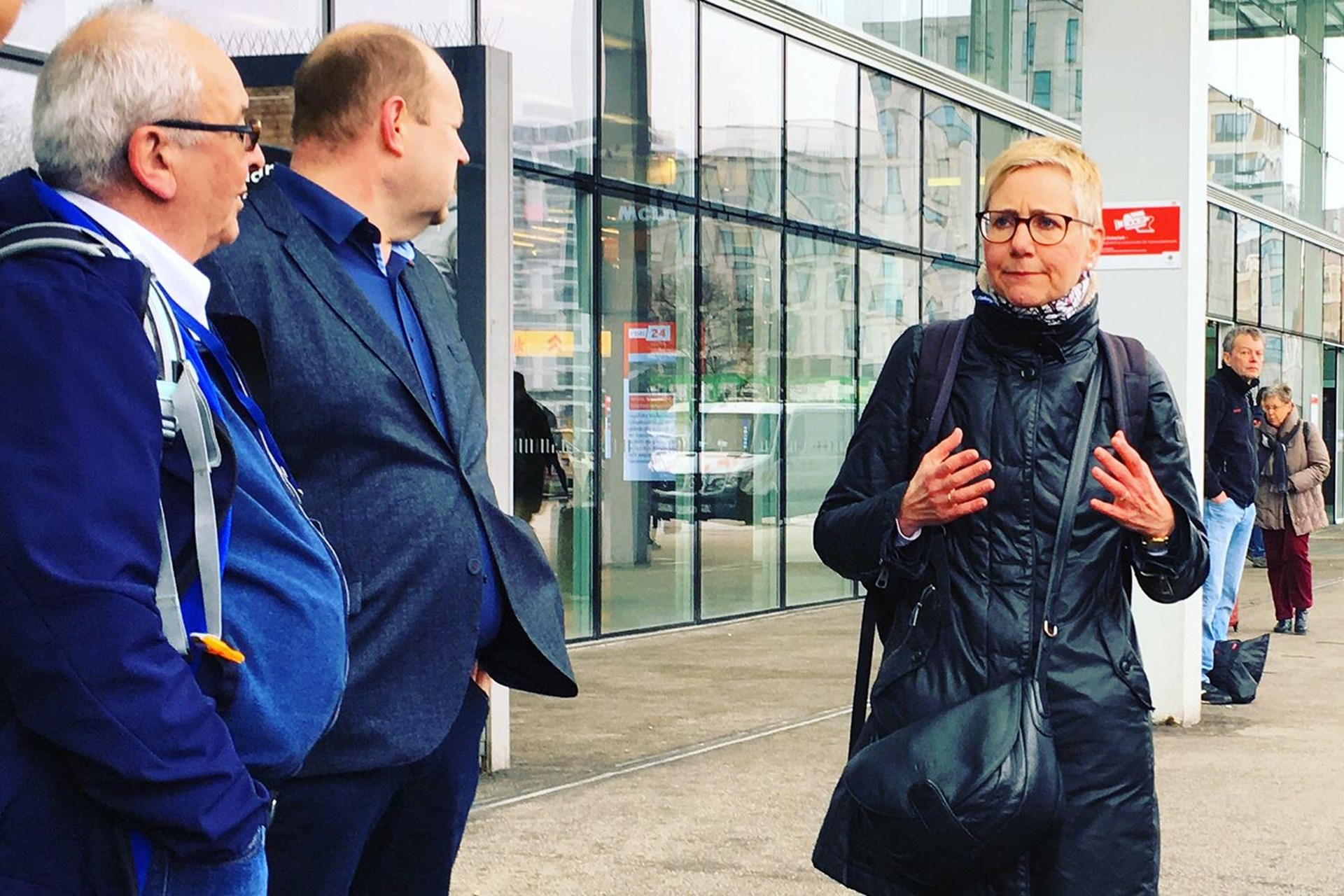Drei Personen stehen vor einem modernen Gebäude. Eine Frau in schwarzer Jacke spricht gestikulierend, während zwei Männer, aufmerksam zuhören. 