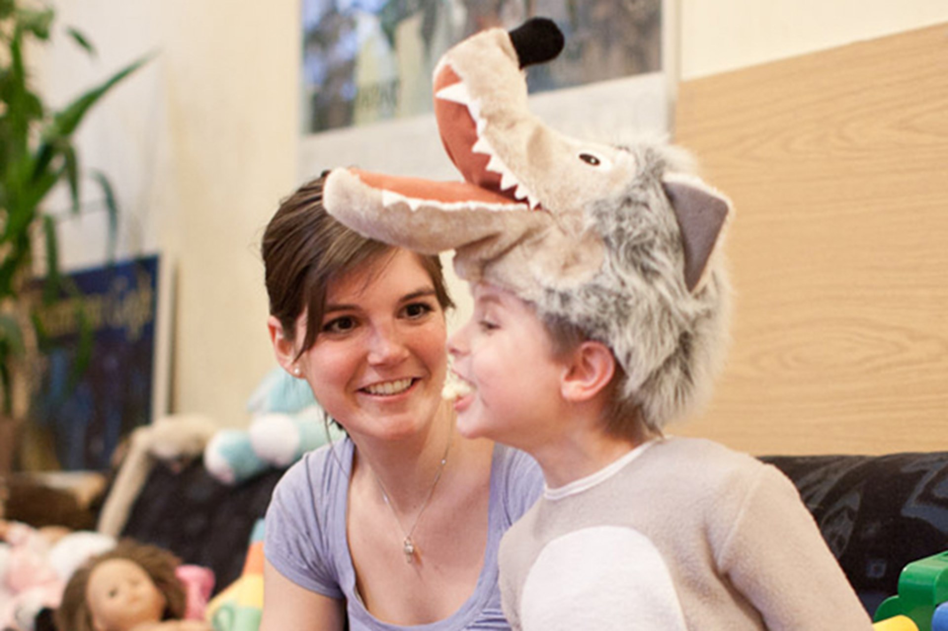 Ein Kind in einem Wolfskostüm mit einer Wolfsmütze lächelt, während eine Frau ihm aufmerksam zusieht. Im Hintergrund sind Spielzeuge und eine Pflanze sichtbar.