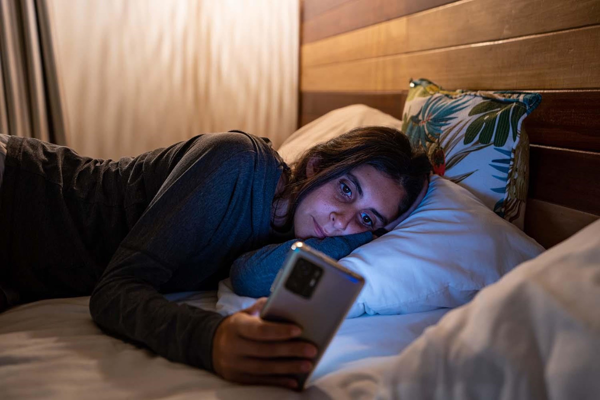 Junge Frau liegt auf einem Bett und schaut auf ihr Smartphone, die Beine angewinkelt und das Gesicht in einen Kissen gebettet, in einem gedämpften, nächtlichen Raum.