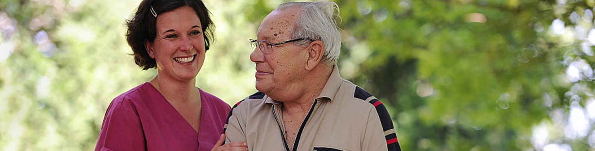 Eine lächelnde Frau in rosa Pflegekleidung steht neben einem älteren Mann mit Brille. Beide wirken freundlich und entspannt, umgeben von grüner Natur.