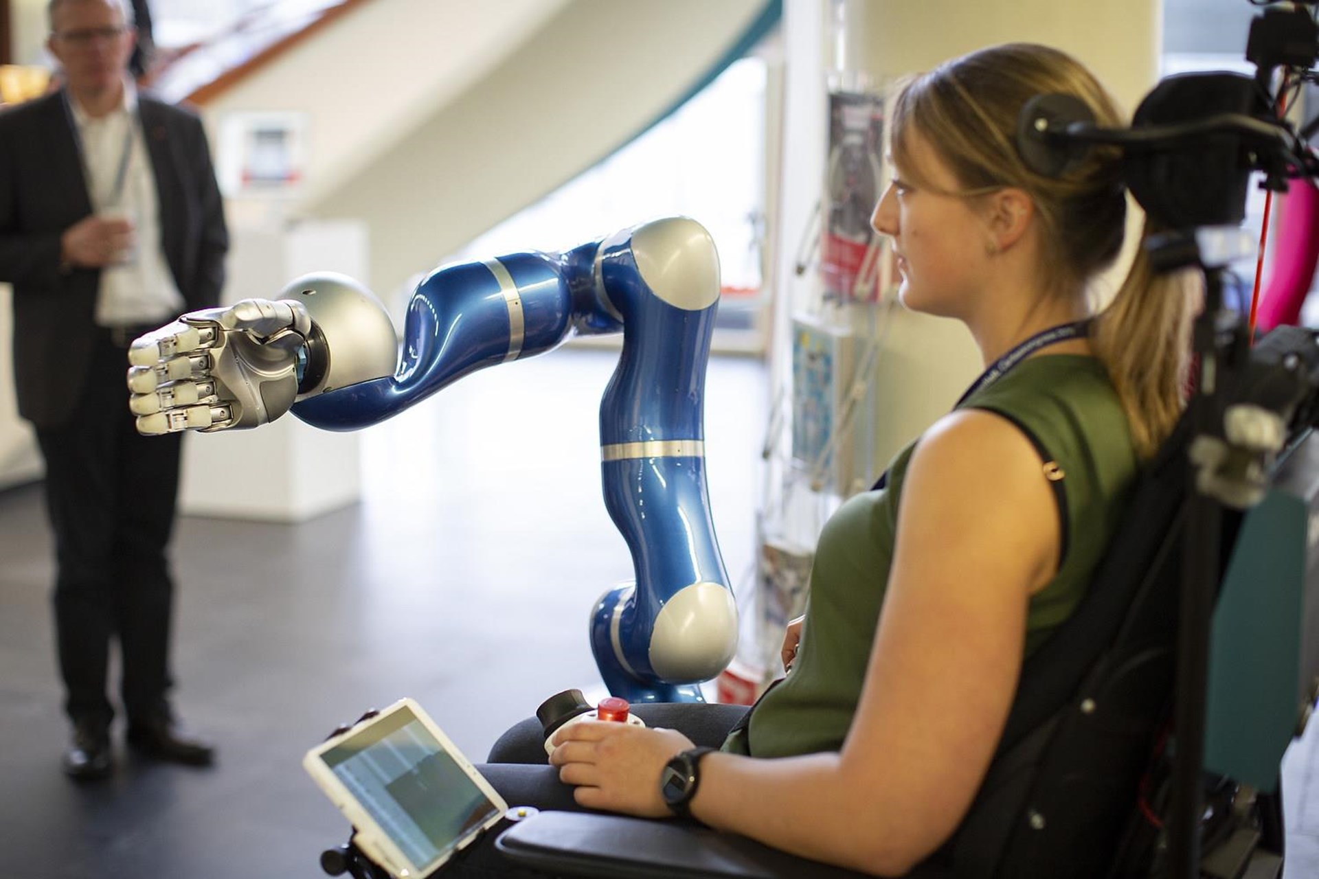 Eine Frau sitzt in einem Rollstuhl und interagiert mit einem blauen Roboterarm. 