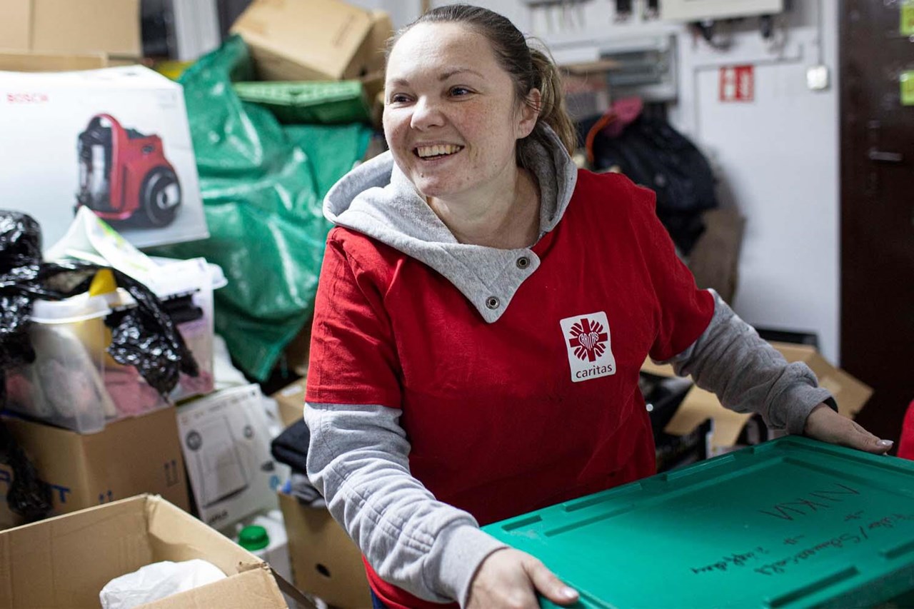 Frau in rotem T-Shirt mit Logo von Caritas, lächelt und trägt eine grüne Kiste in einem Raum voller Kartons und Müll.