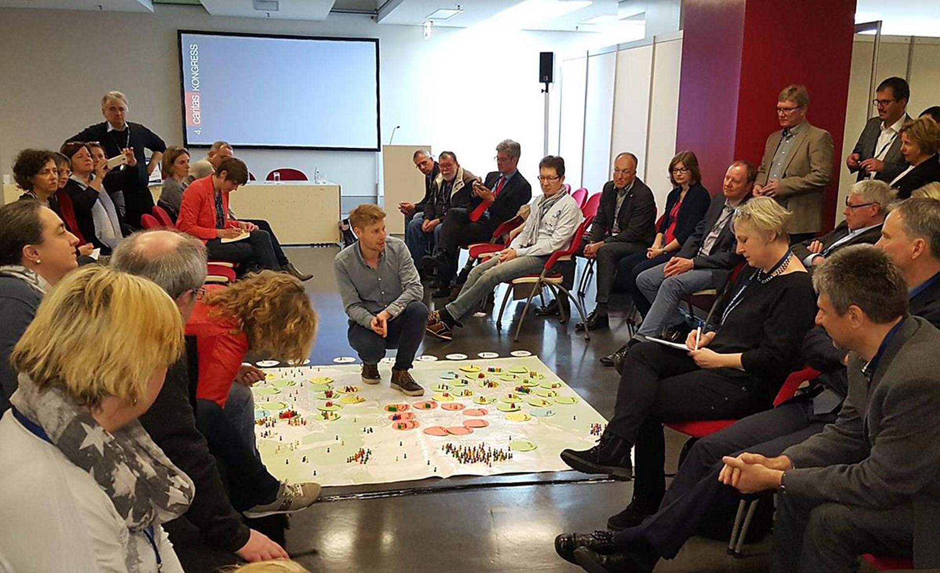 Gruppendiskussion in einem Konferenzraum mit Teilnehmern, die um ein großes, interaktives Plakat sitzen. Ein Moderator erklärt die Inhalte.