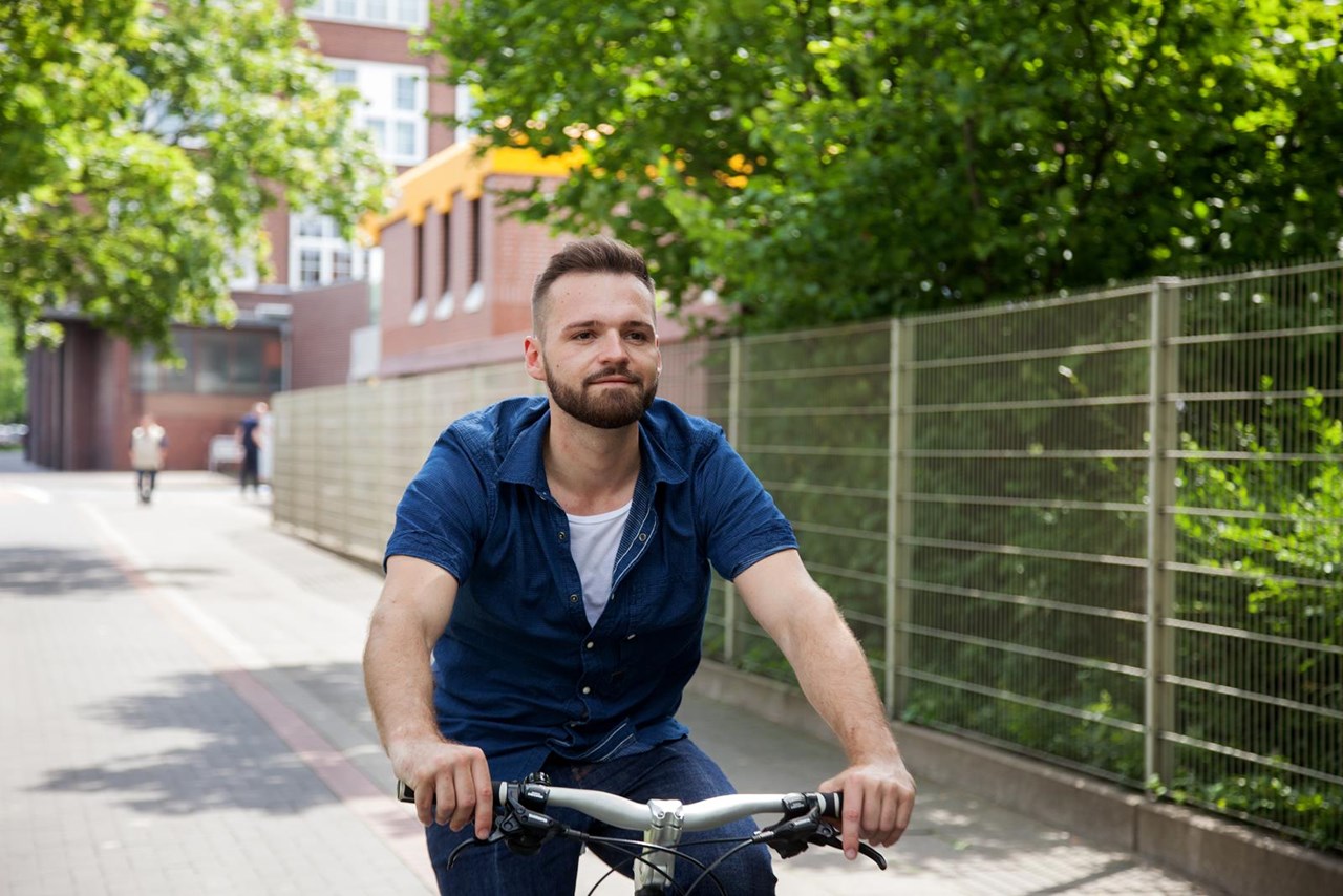 Mann mit Bart fährt auf einem Fahrrad in einer städtischen Umgebung, umgeben von Bäumen und einem Zaun.