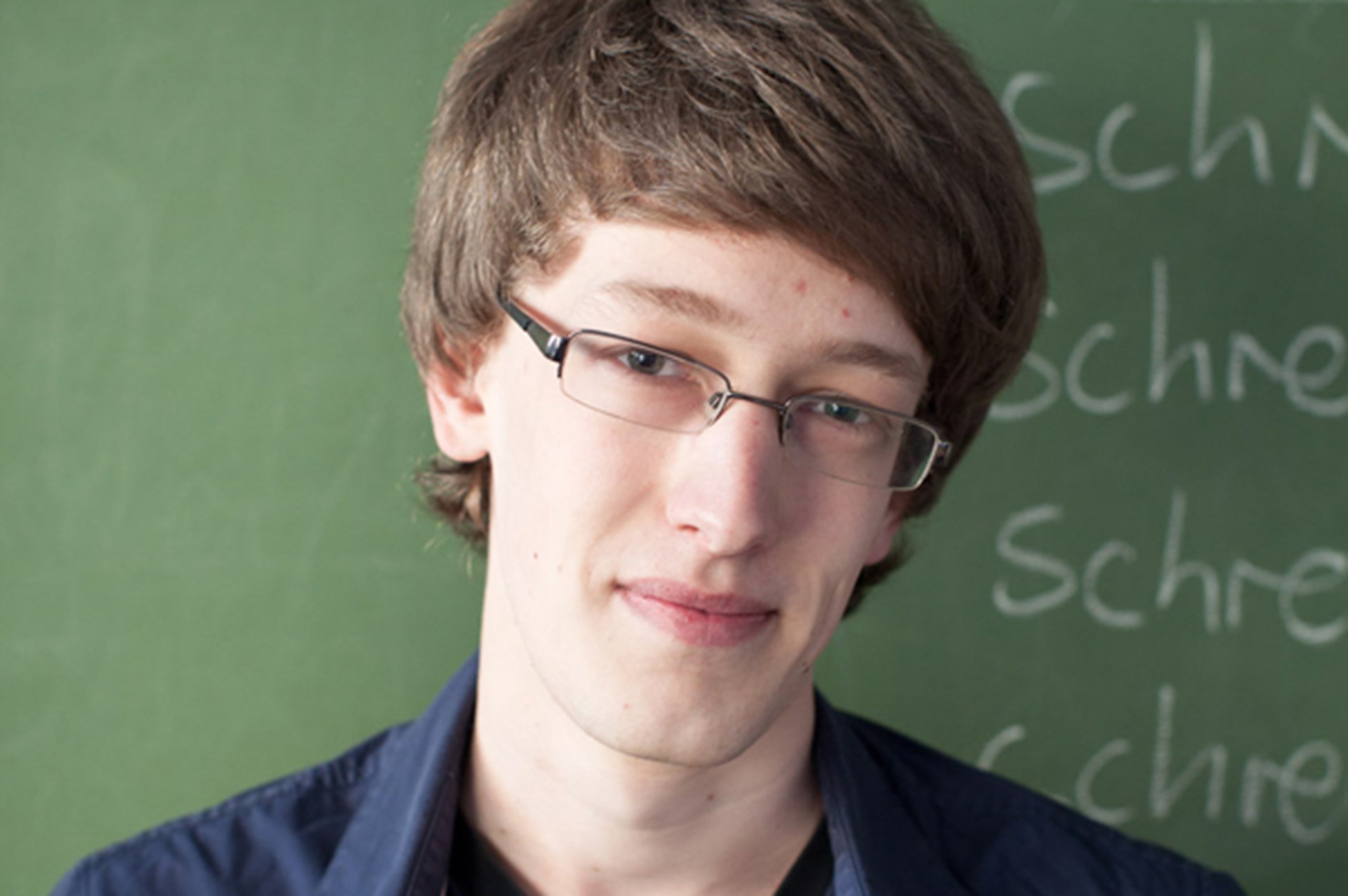 Junger Mann mit Brille und lockigem Haar vor einer Tafel mit handschriftlichen Notizen.