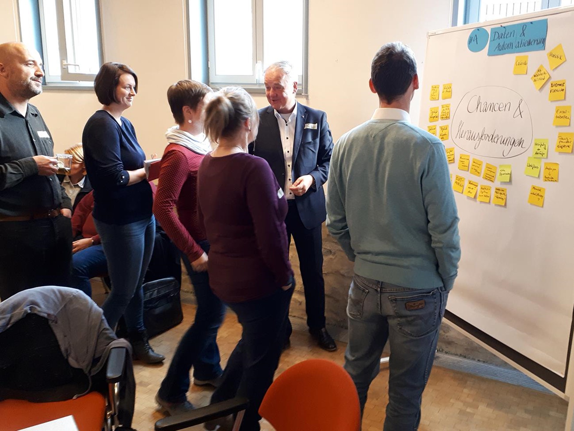 Gruppendiskussion in einem Seminarraum, Teilnehmer stehen um ein Flipchart mit Haftnotizen. Personen in lockerer Kleidung, einige lächeln und interagieren.