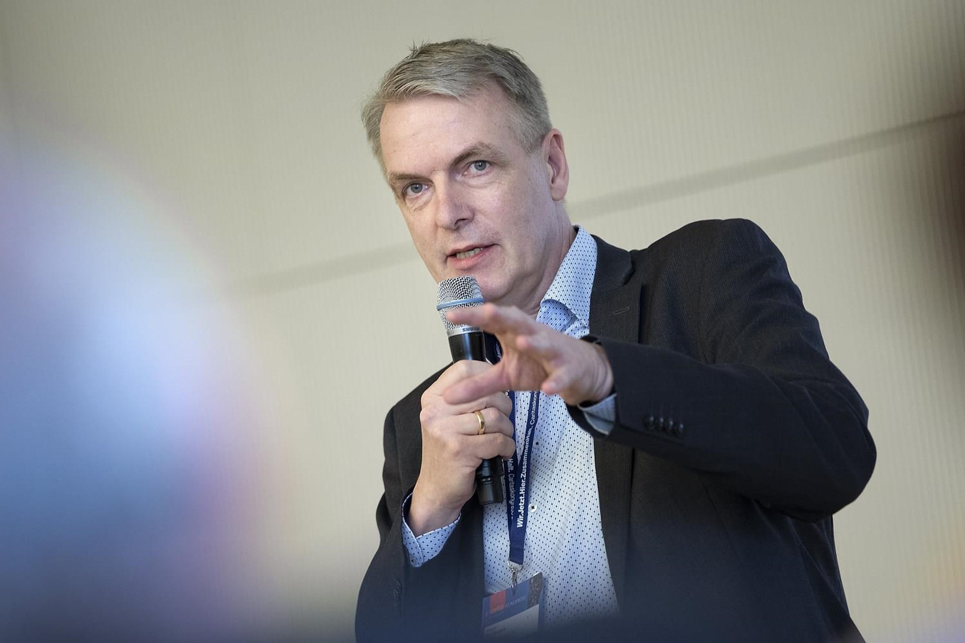 Mann mit Mikrofon spricht auf einer Konferenz, trägt einen dunklen Anzug und ein helles Hemd, gestikuliert mit der Hand.