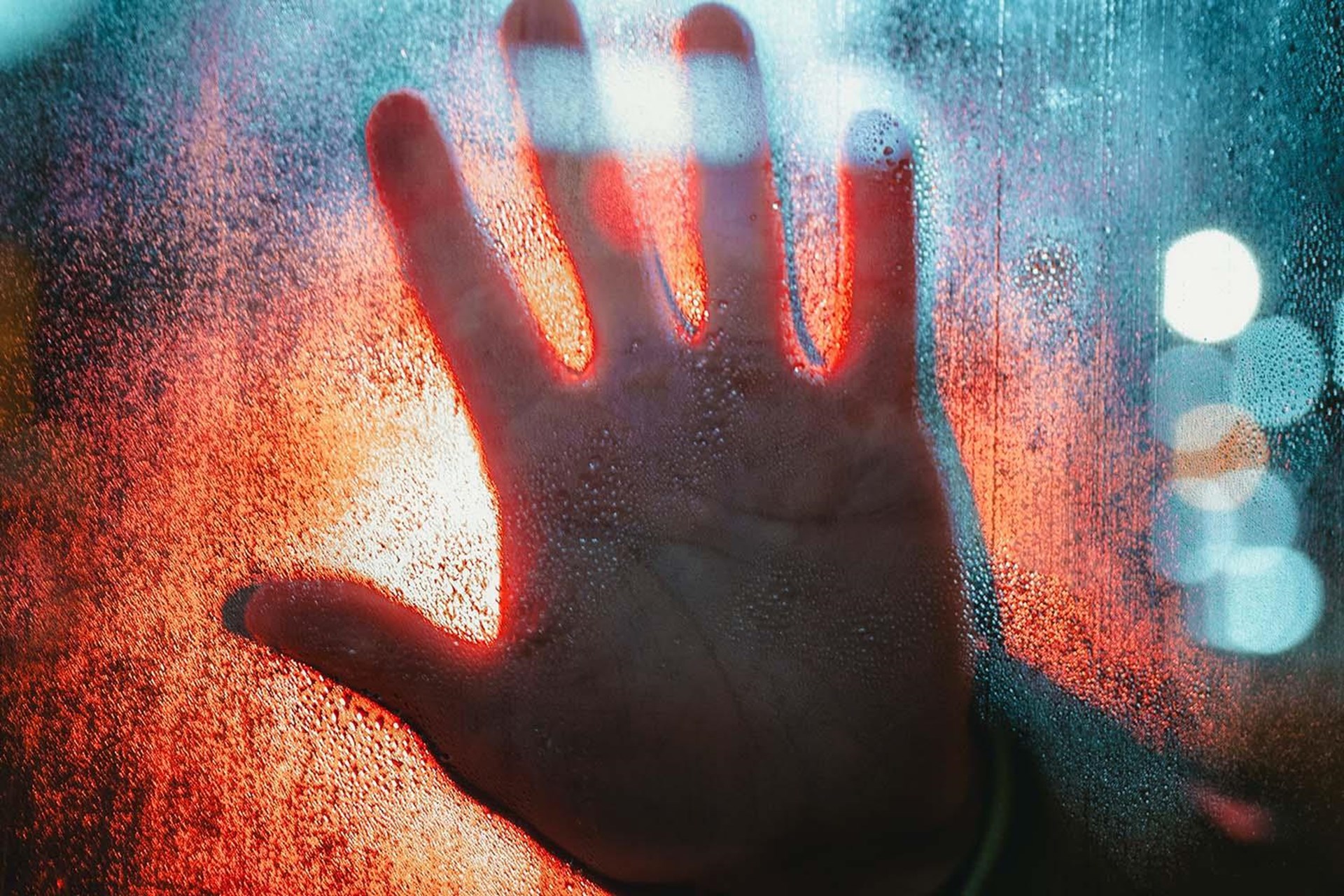 Eine Hand drückt von außen an eine Glasscheibe, auf der Wassertropfen sind. Im Hintergrund scheint rotes Licht.